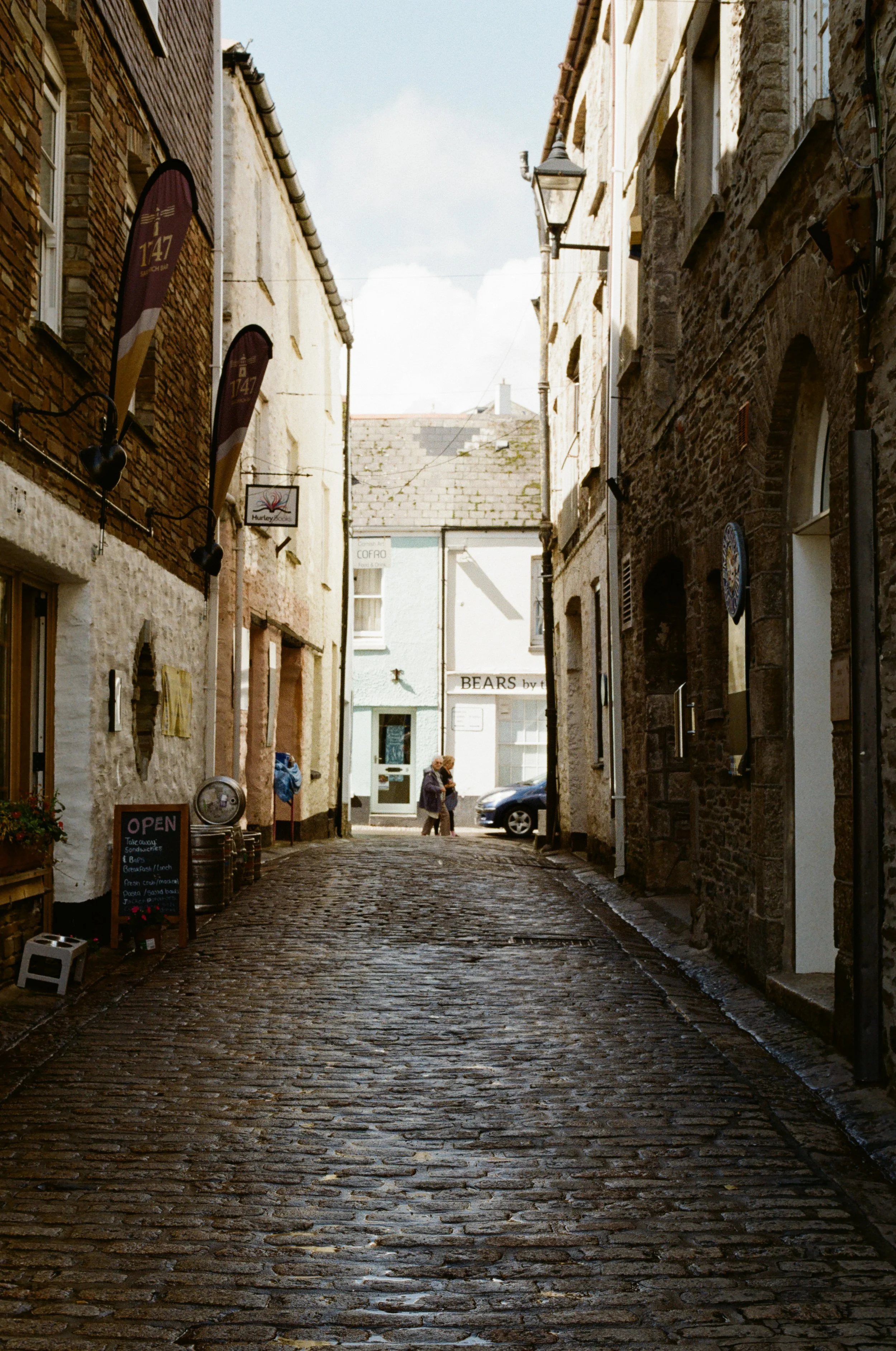 A narrow cobblestone street in a historic town with stone and brick buildings on either side, a few pedestrians walking, and small shops and signs visible.