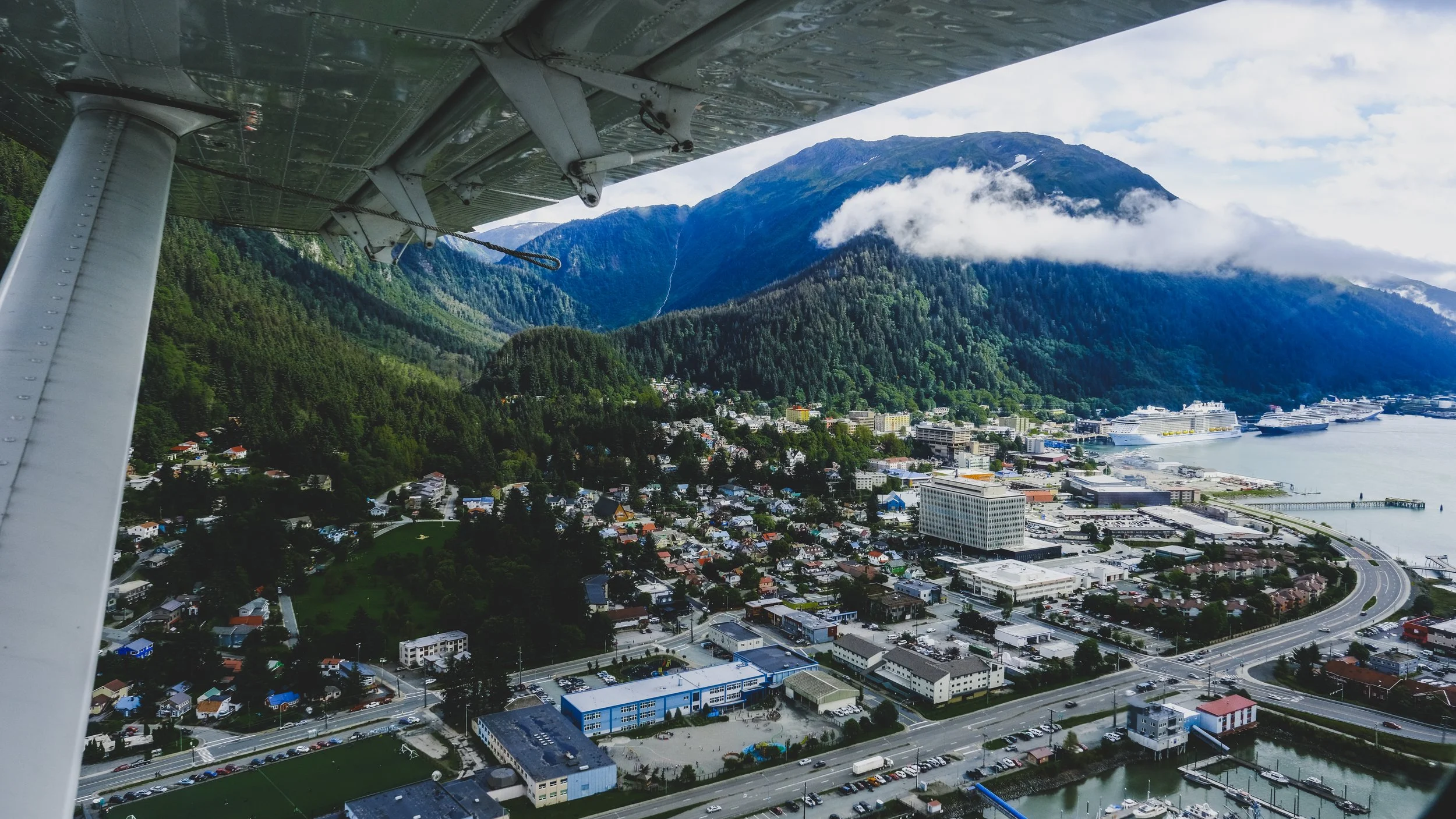 Aerial view of a coastal town with mountains in the background, cruise ships docked at the harbor, and a small airplane wing visible in the foreground.