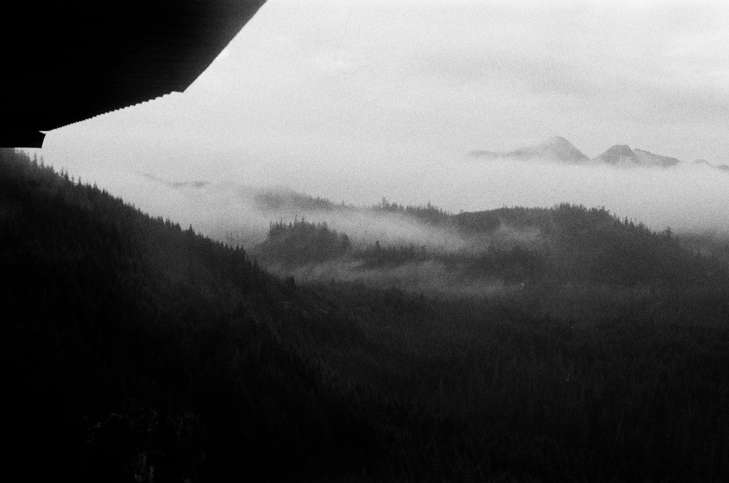 Black and white photograph of a mountain landscape with fog and distant peaks, taken from under an overhanging roof.
