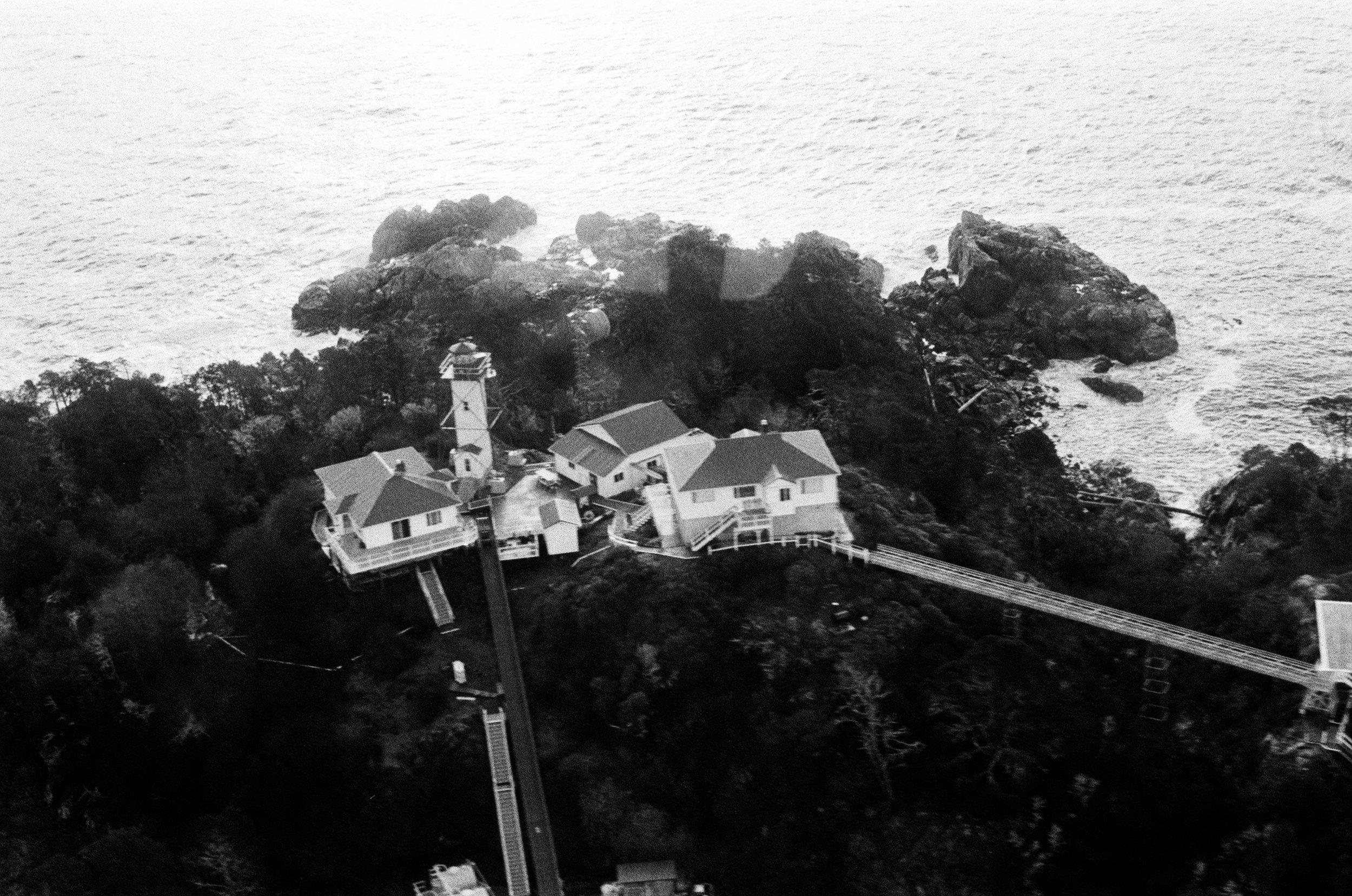 A black and white aerial view of a coastal house with a lighthouse, connected by a long walkway, near rocky shoreline with trees and ocean.