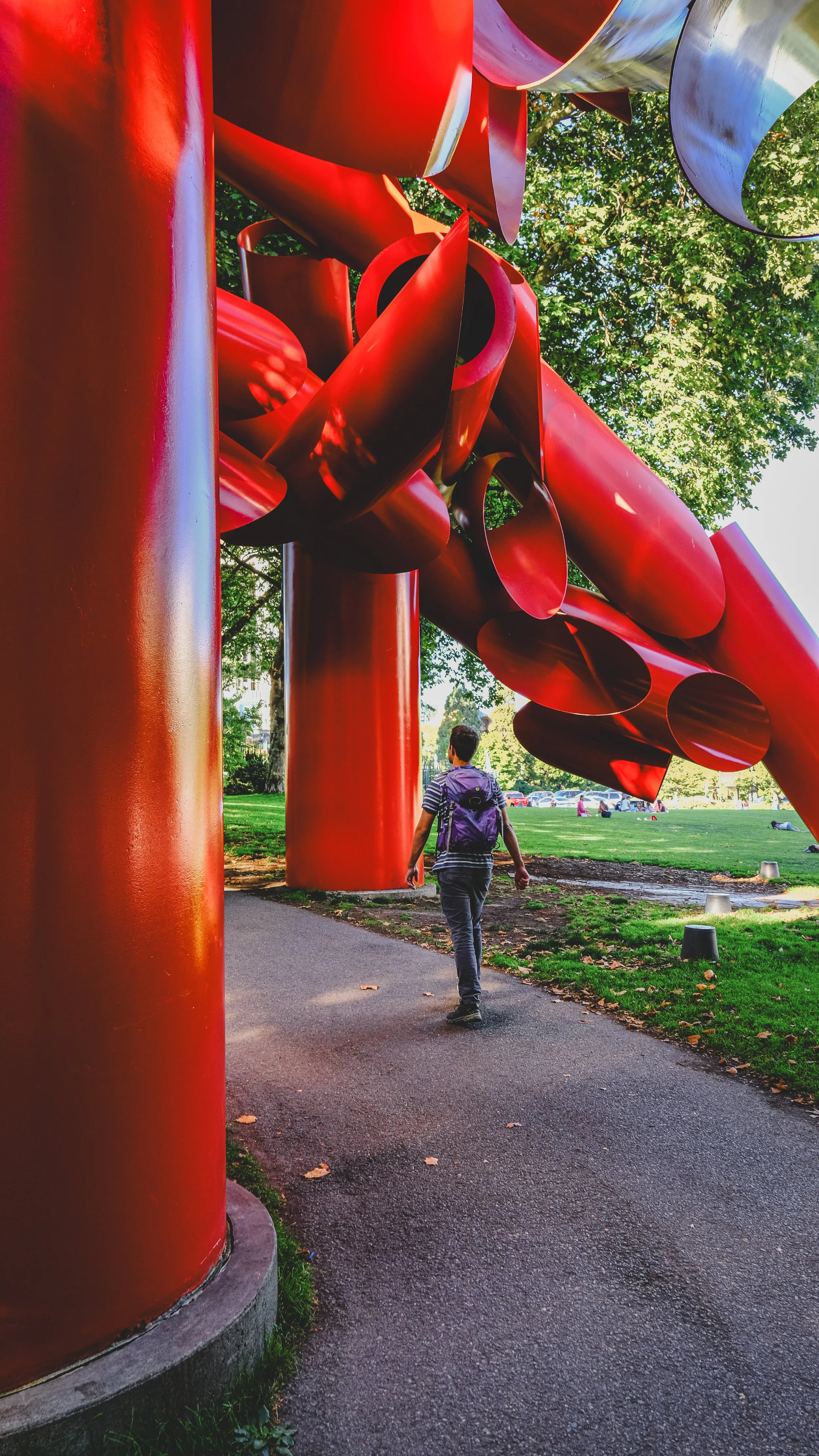 A person walking along a paved path near a large, modern red sculpture in a park setting, with trees and open grassy area in the background.