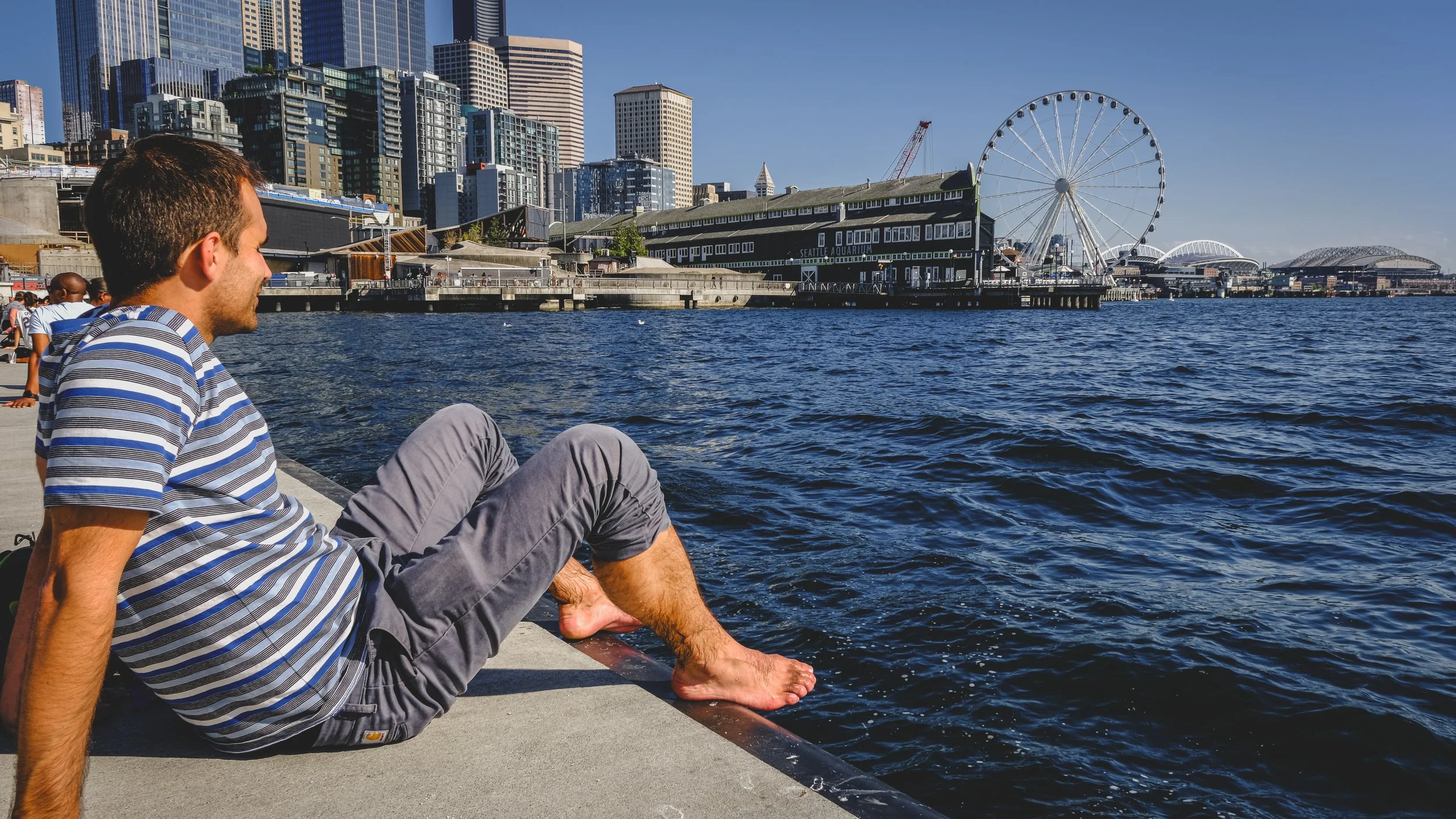 Man in a striped shirt sitting on dock with feet in water, overlooking Seattle waterfront with city buildings and Ferris wheel.
