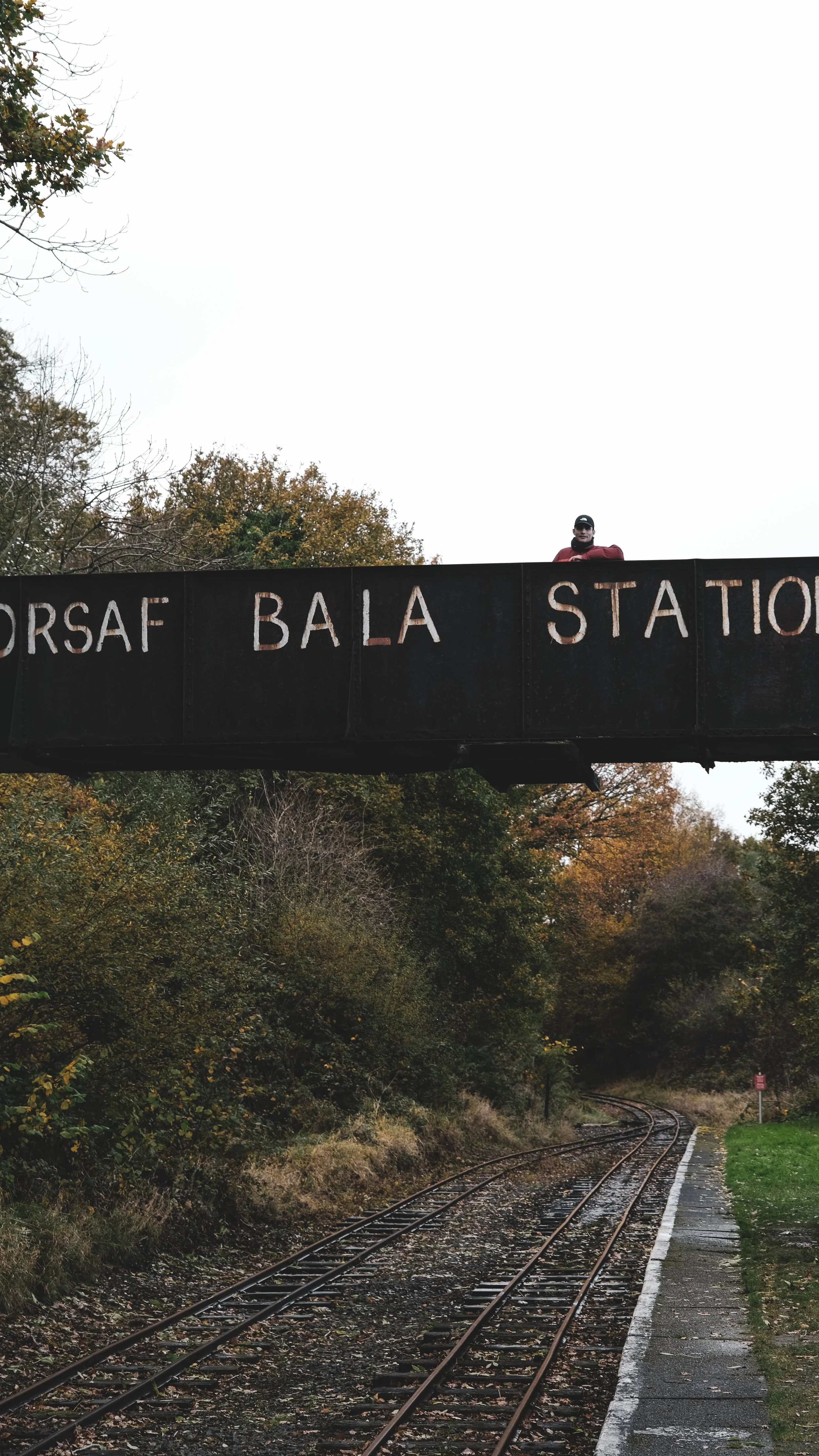 A railway track running through a wooded area with trees showing autumn colors, and a bridge labeled 'GORSF BALA STATION' with a person standing on it.