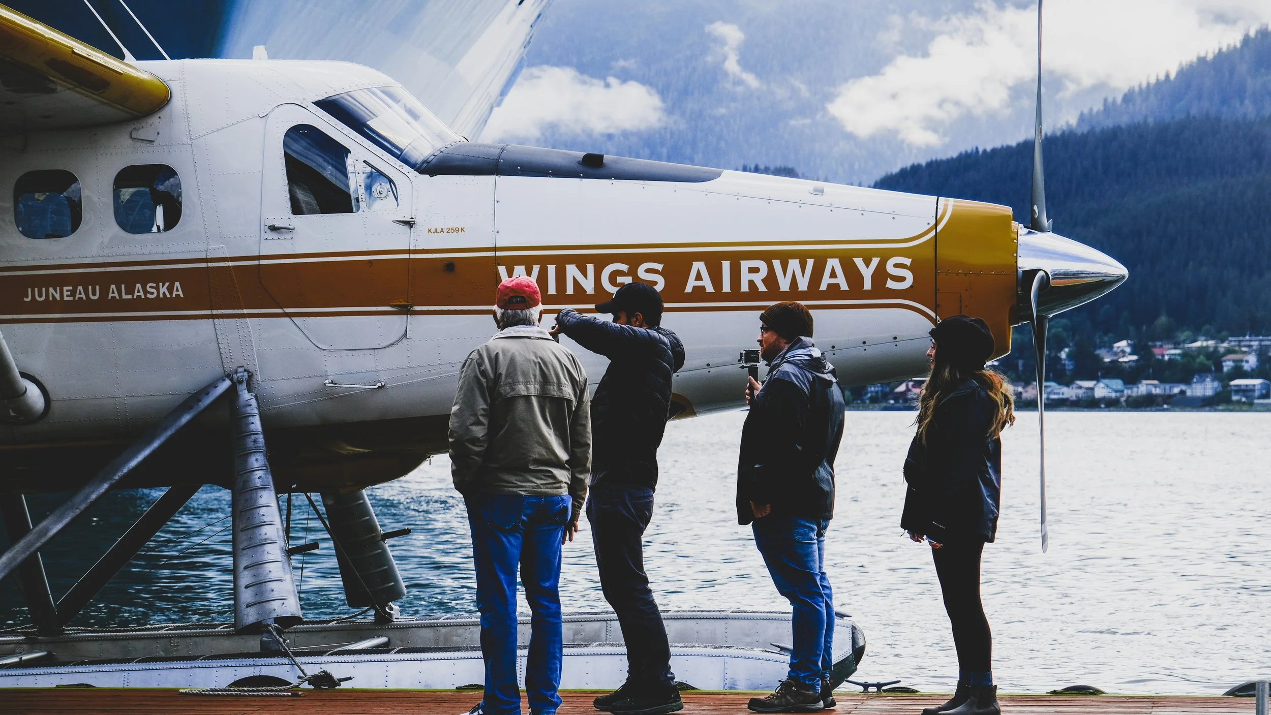 Four people standing on a dock near a floatplane with 'WINGS AIRWAYS' and 'JUNEAU ALASKA' written on it, with a lake and forested mountains in the background.