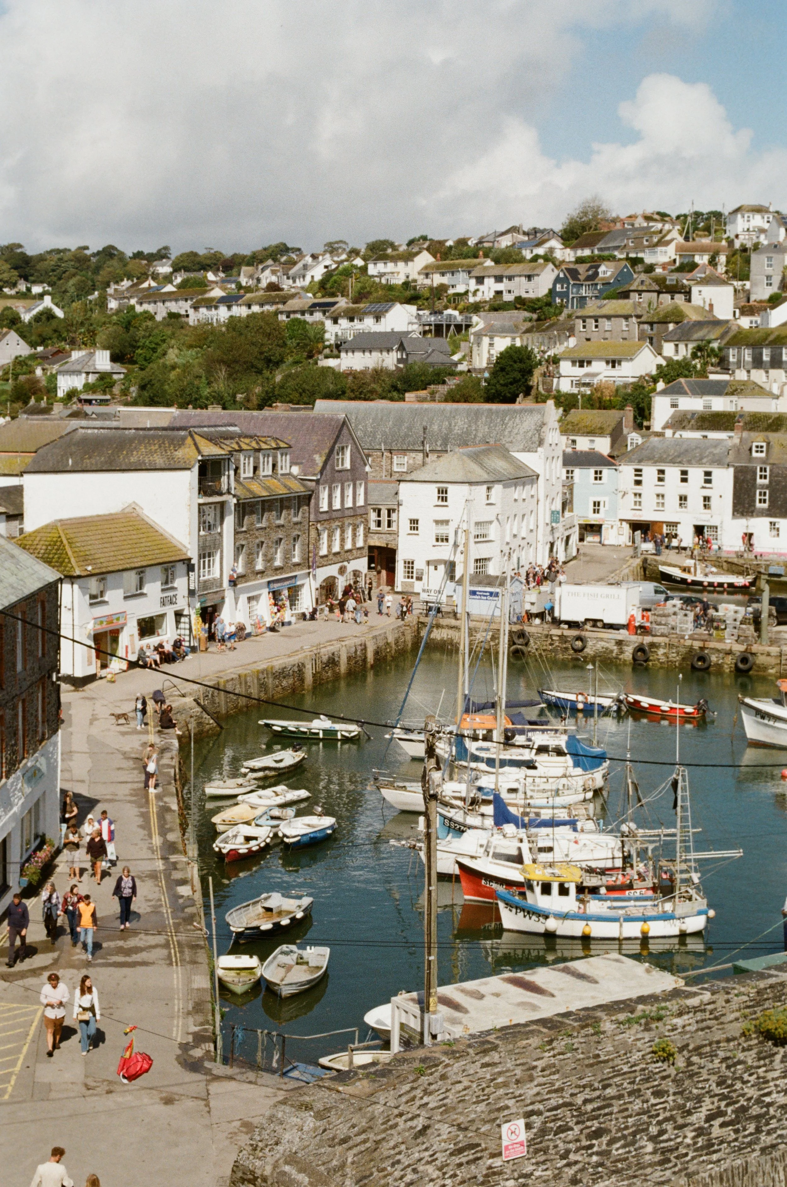 A coastal town with a harbor filled with boats, surrounded by old buildings and houses on a hillside under a partly cloudy sky.
