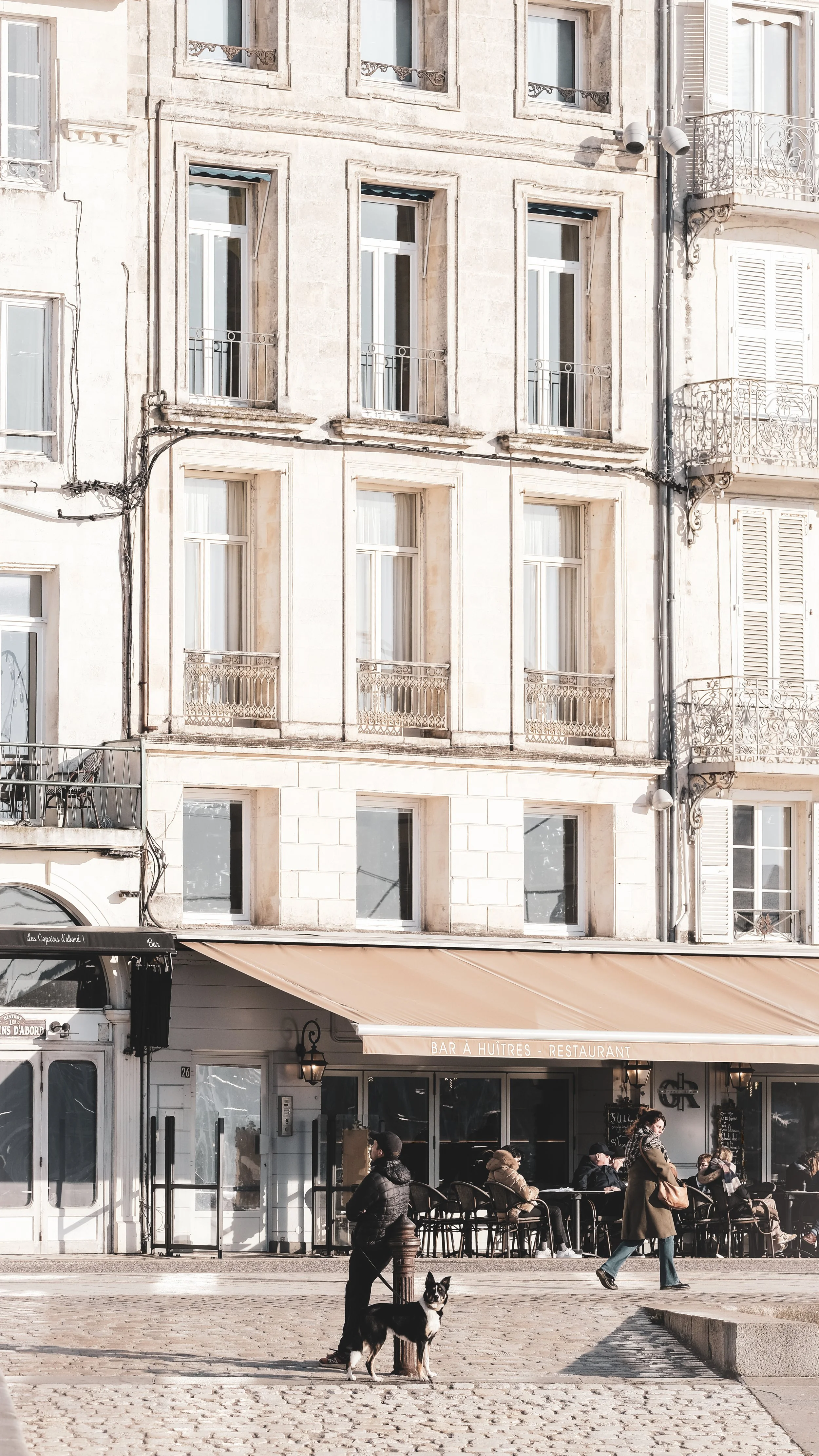 Scene of a city street with a café and restaurant called 'Bar à Huîtres - Restaurant' with outdoor seating areas. People are sitting outside, and a woman with a dog is standing nearby. The building behind has multiple floors with windows and small balconies, featuring beige stone facades and decorative black wrought-iron railings.