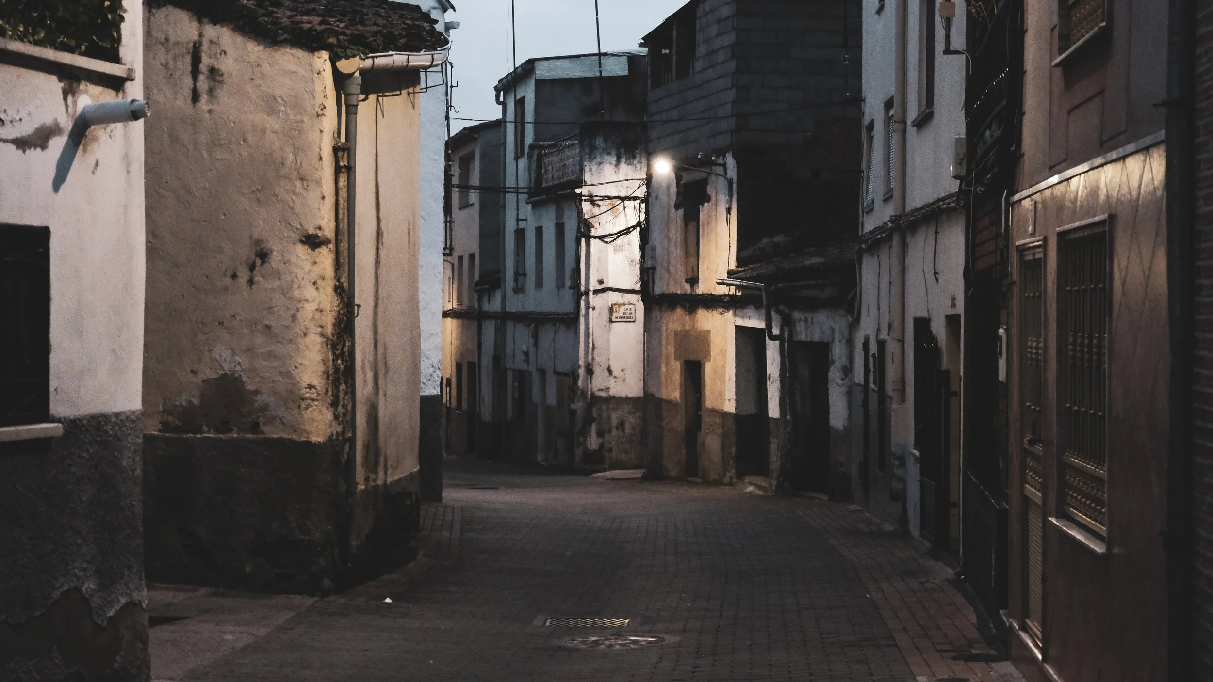 A narrow, empty alleyway at dusk with old, weathered buildings on both sides, showing peeling paint, barred windows, and dim lighting.