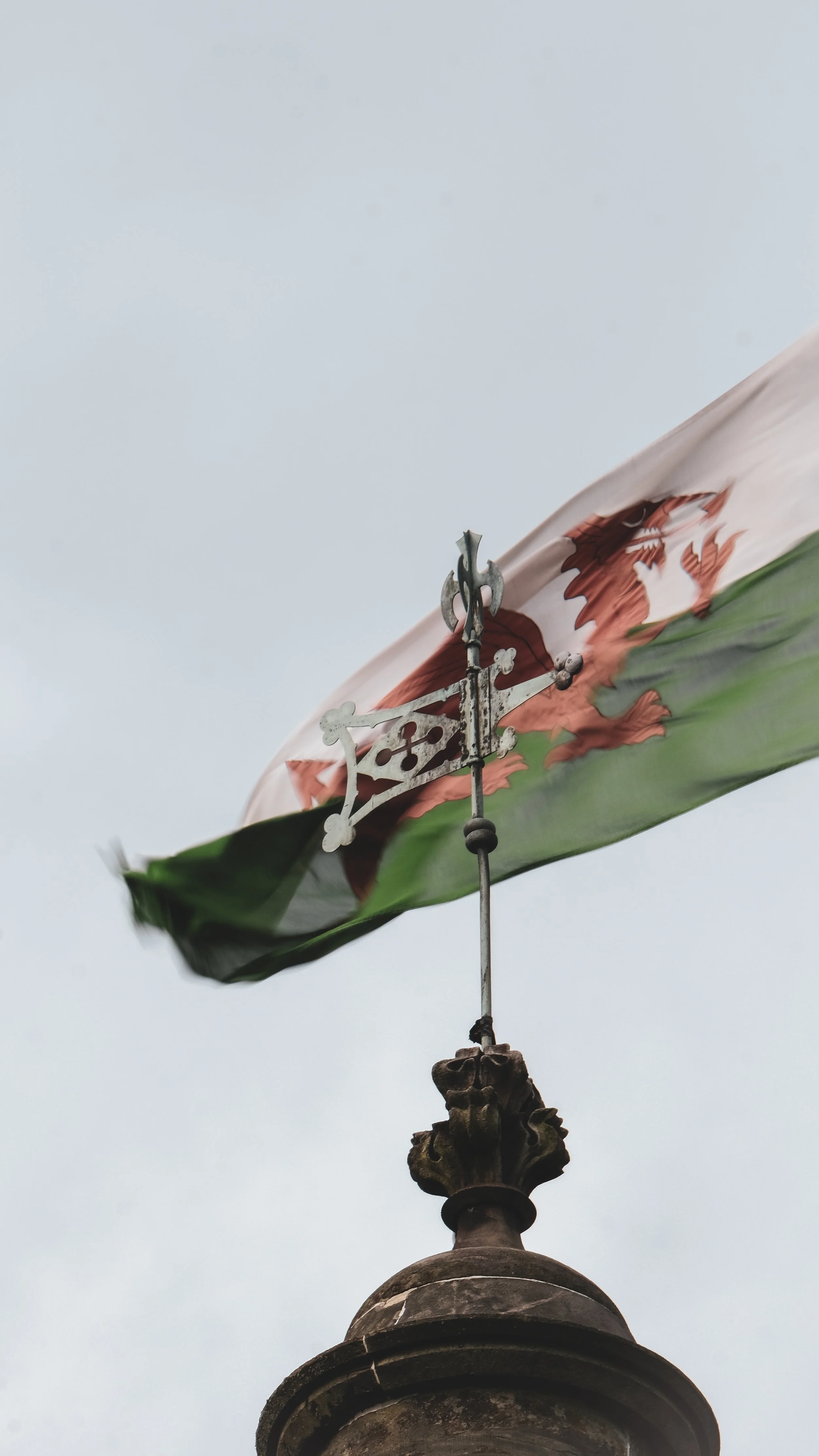 Close-up of a Welsh flag with the red Welsh dragon, mounted on a pole with decorative metalwork, against an overcast sky.