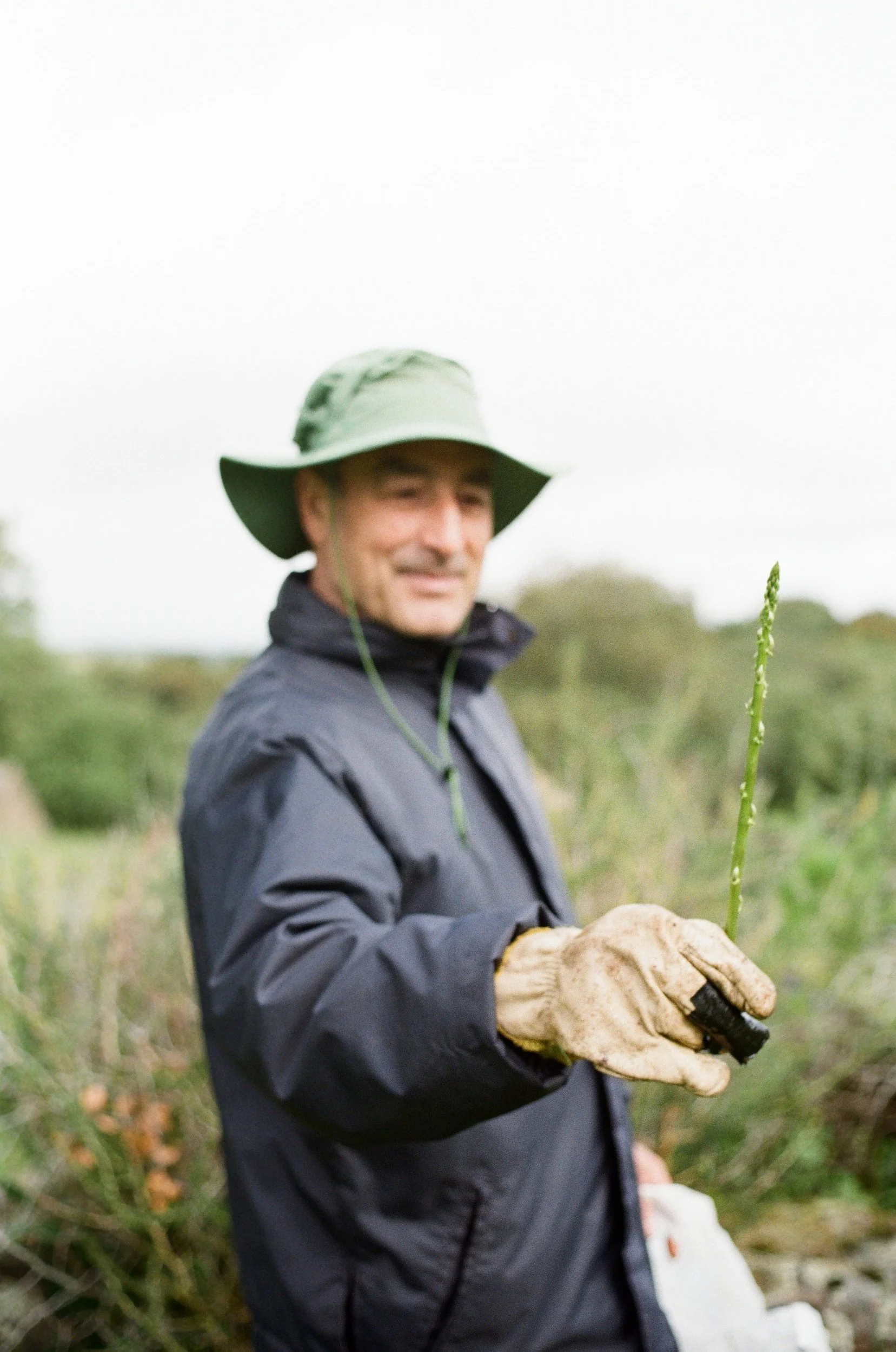 A man wearing a wide-brimmed hat, a dark jacket, and gloves holding a green plant in an outdoor setting with greenery in the background.