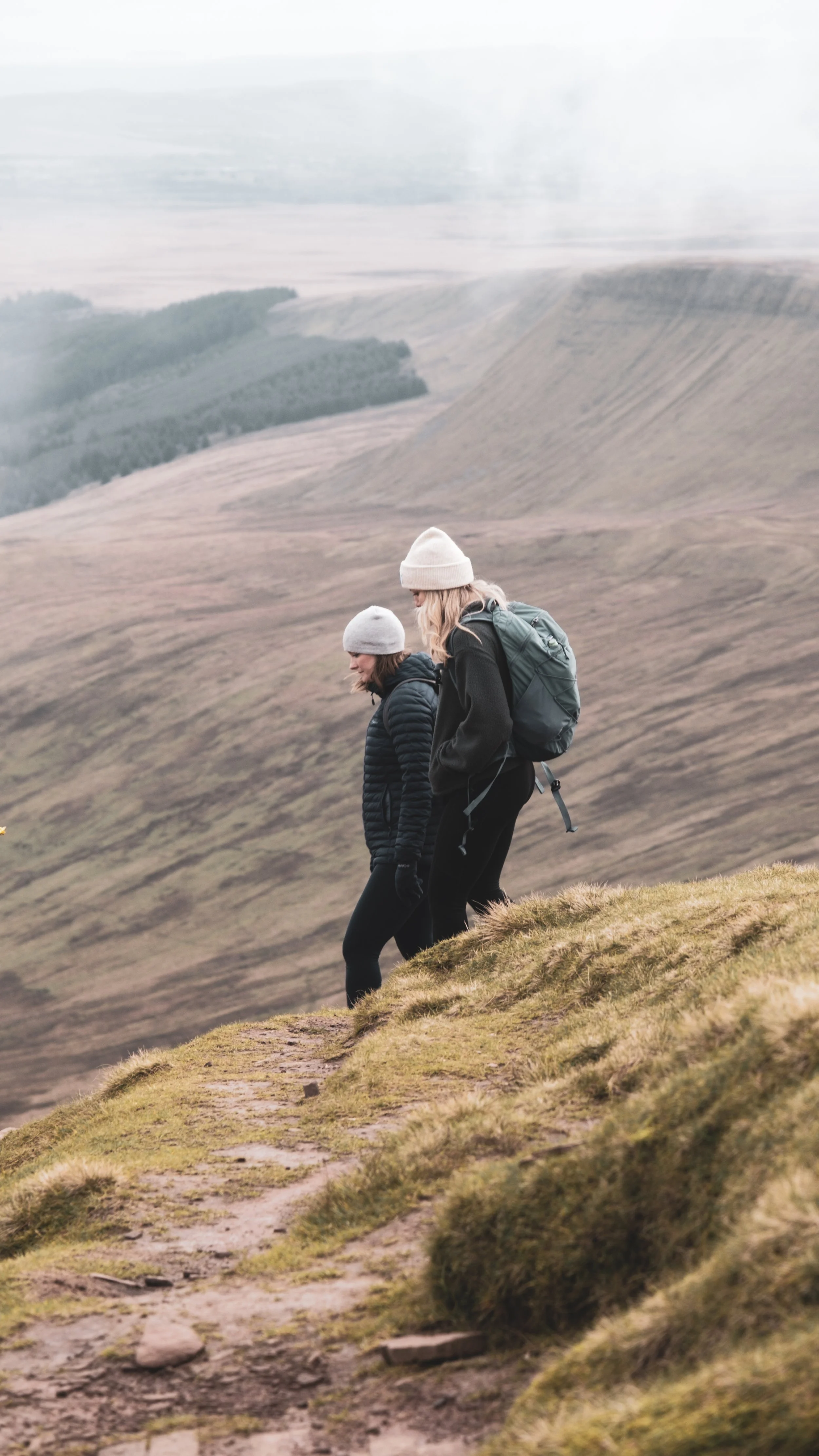Two women hiking on a hill overlooking a valley with rolling hills and sparse vegetation.