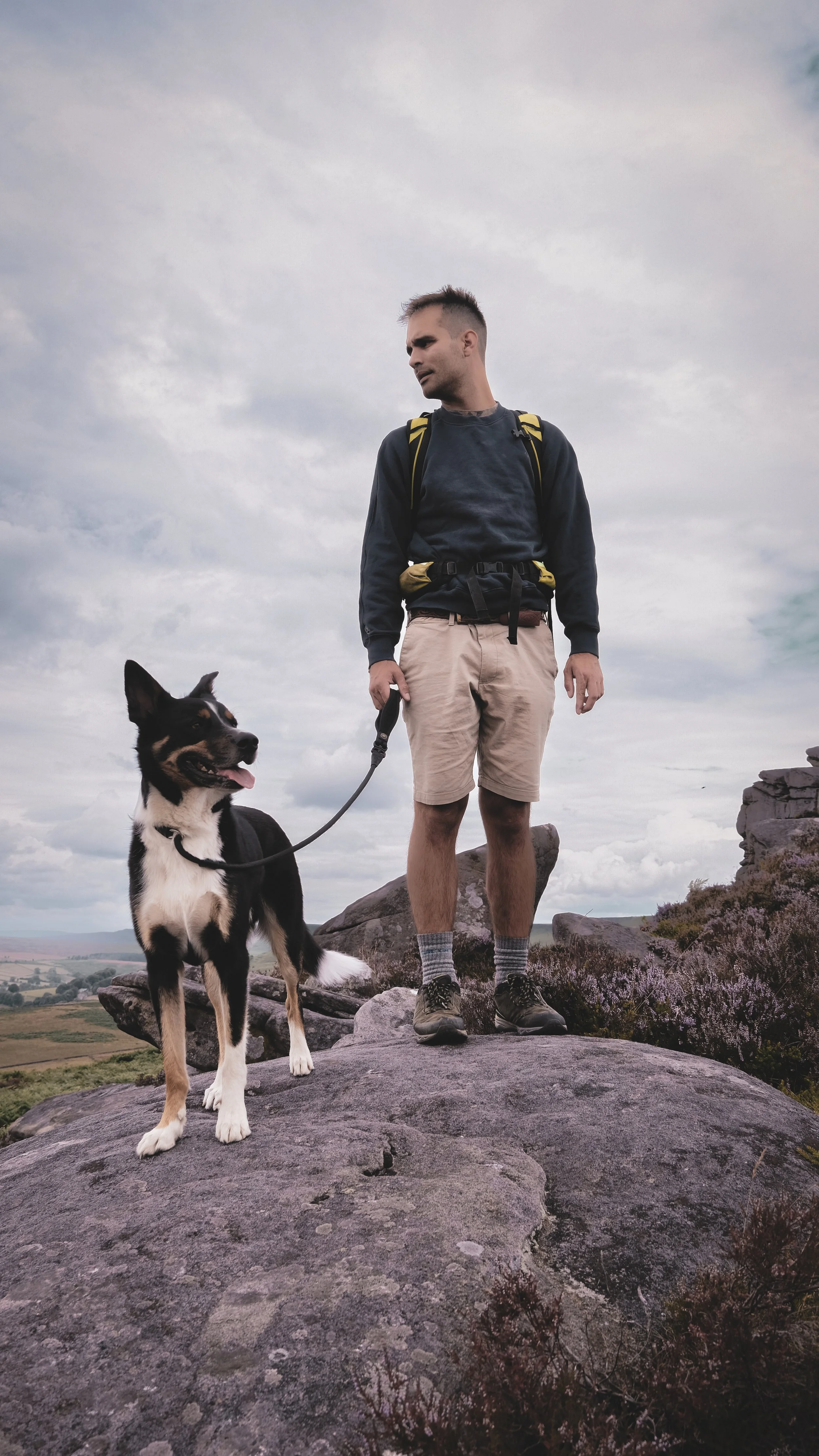 A man with a backpack standing on a rock with his tricolor dog during a hike or outdoor adventure.