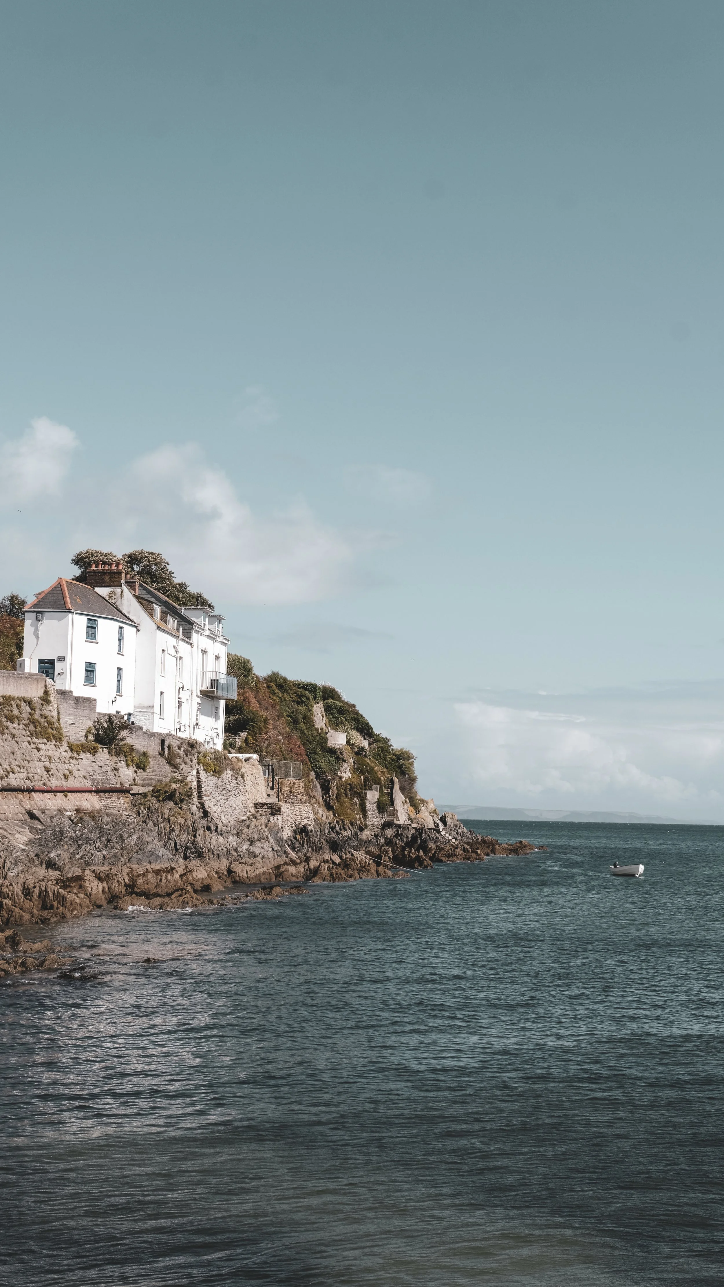 A white house situated on a rocky hill overlooking the ocean with a small boat in the water.