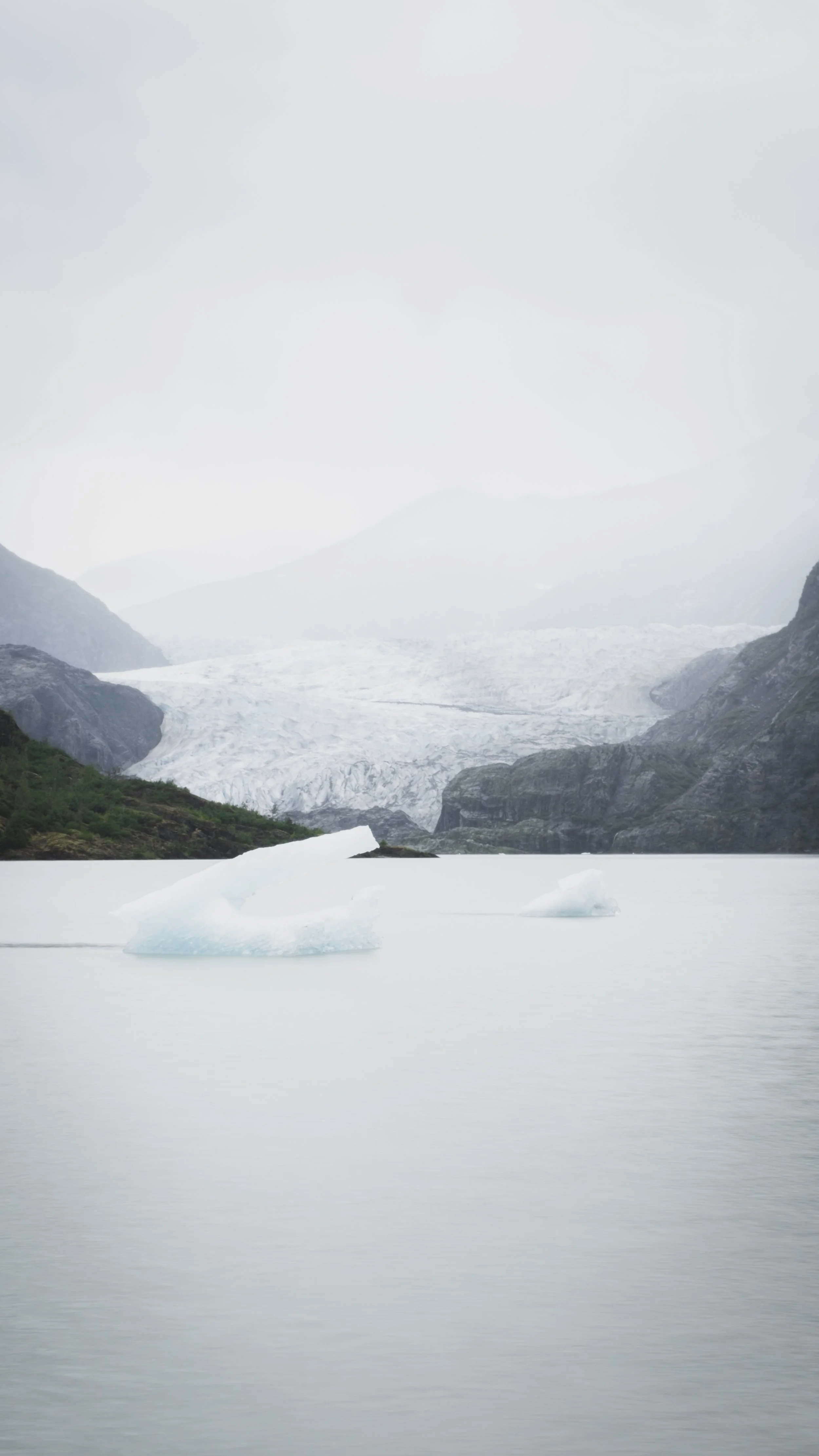 A cold, overcast landscape featuring icy water with icebergs, rocky mountains, and a glacier in the background.