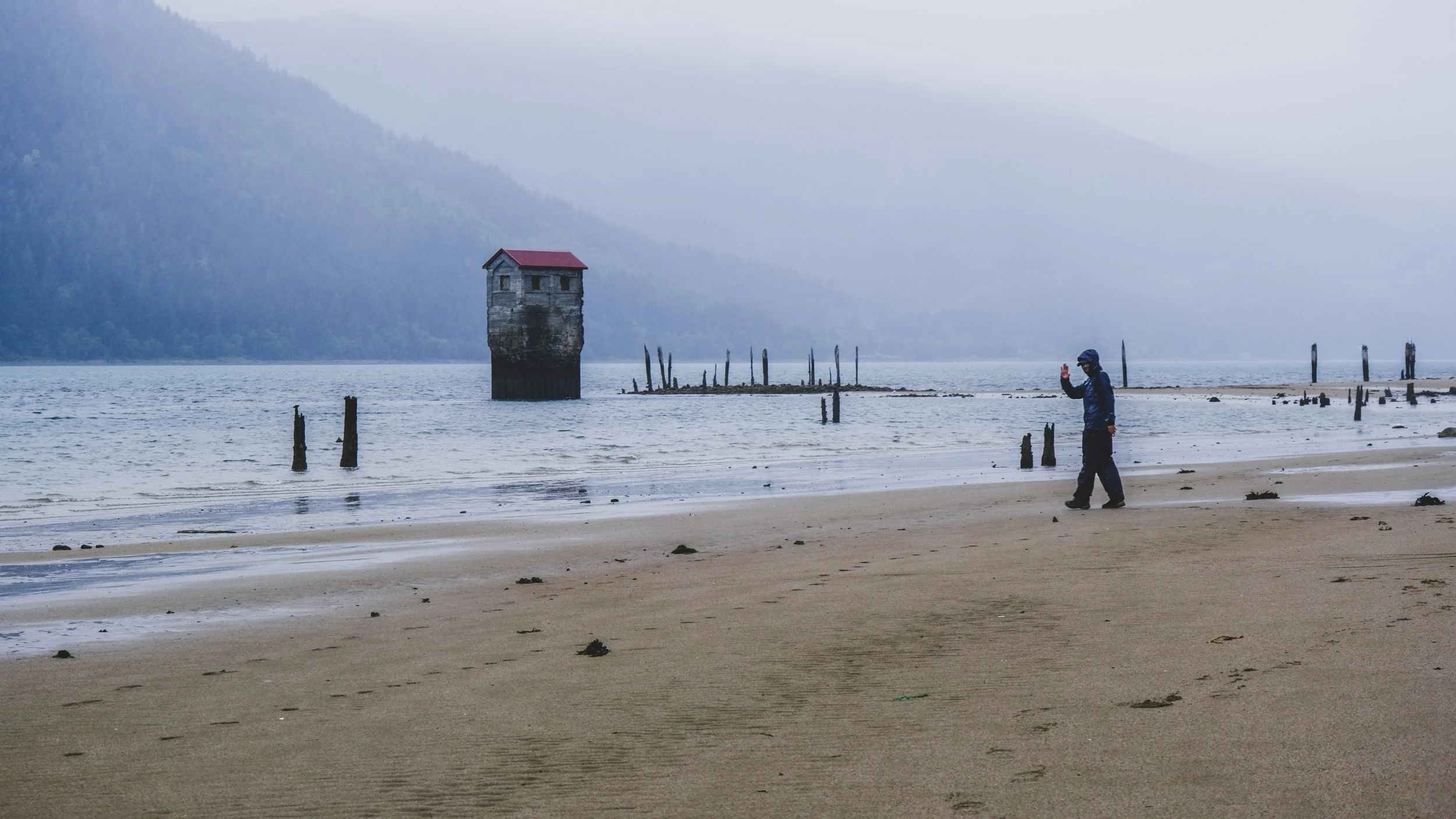 A person in a dark jacket and cap walking on a sandy beach, taking a photo with a smartphone, near old wooden pilings and a small house on stilts in the water against a backdrop of misty mountains.