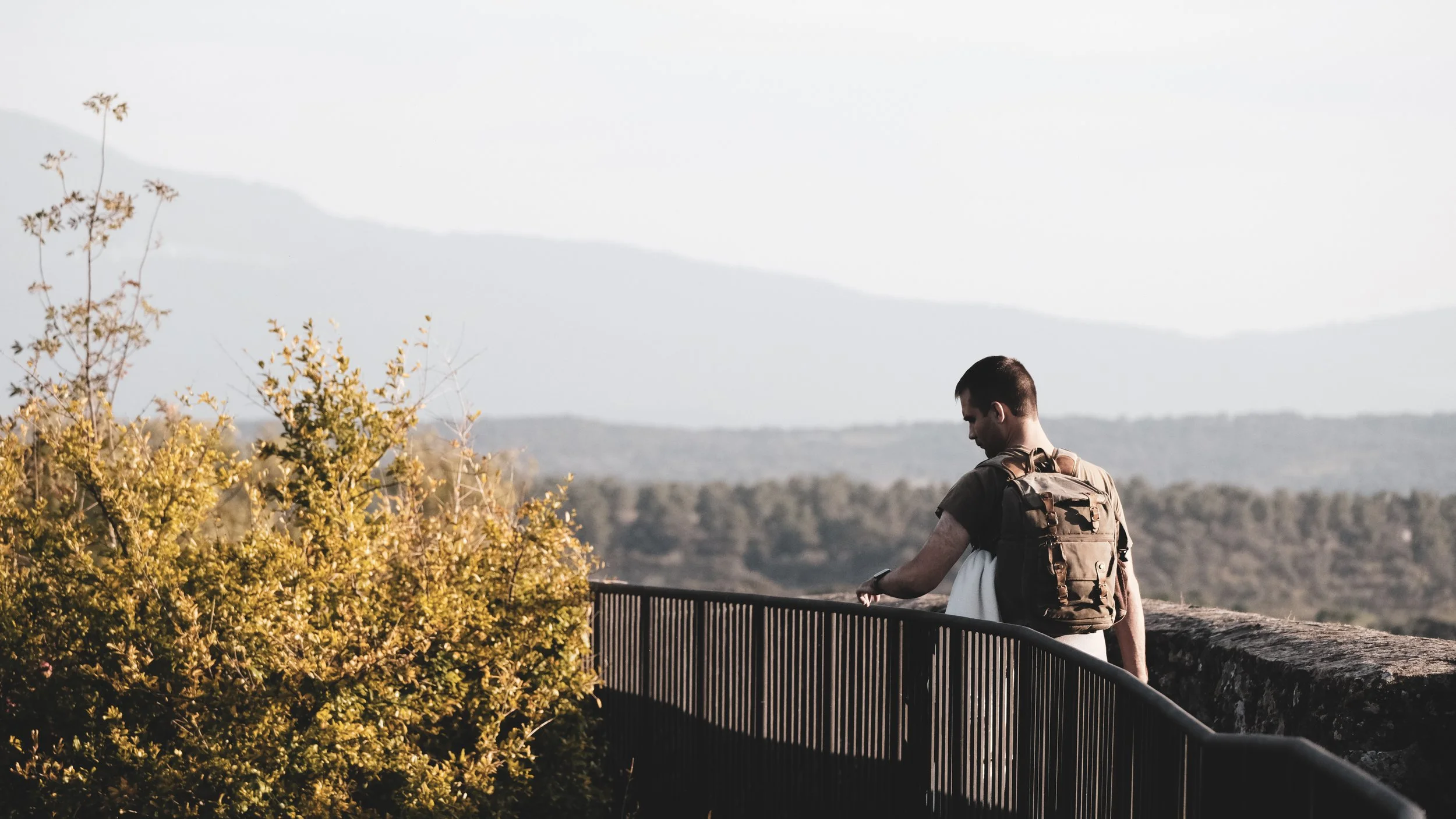 A man with a backpack looking down at a wristwatch on his arm beside a railing, with a scenic landscape of trees and mountains in the background.