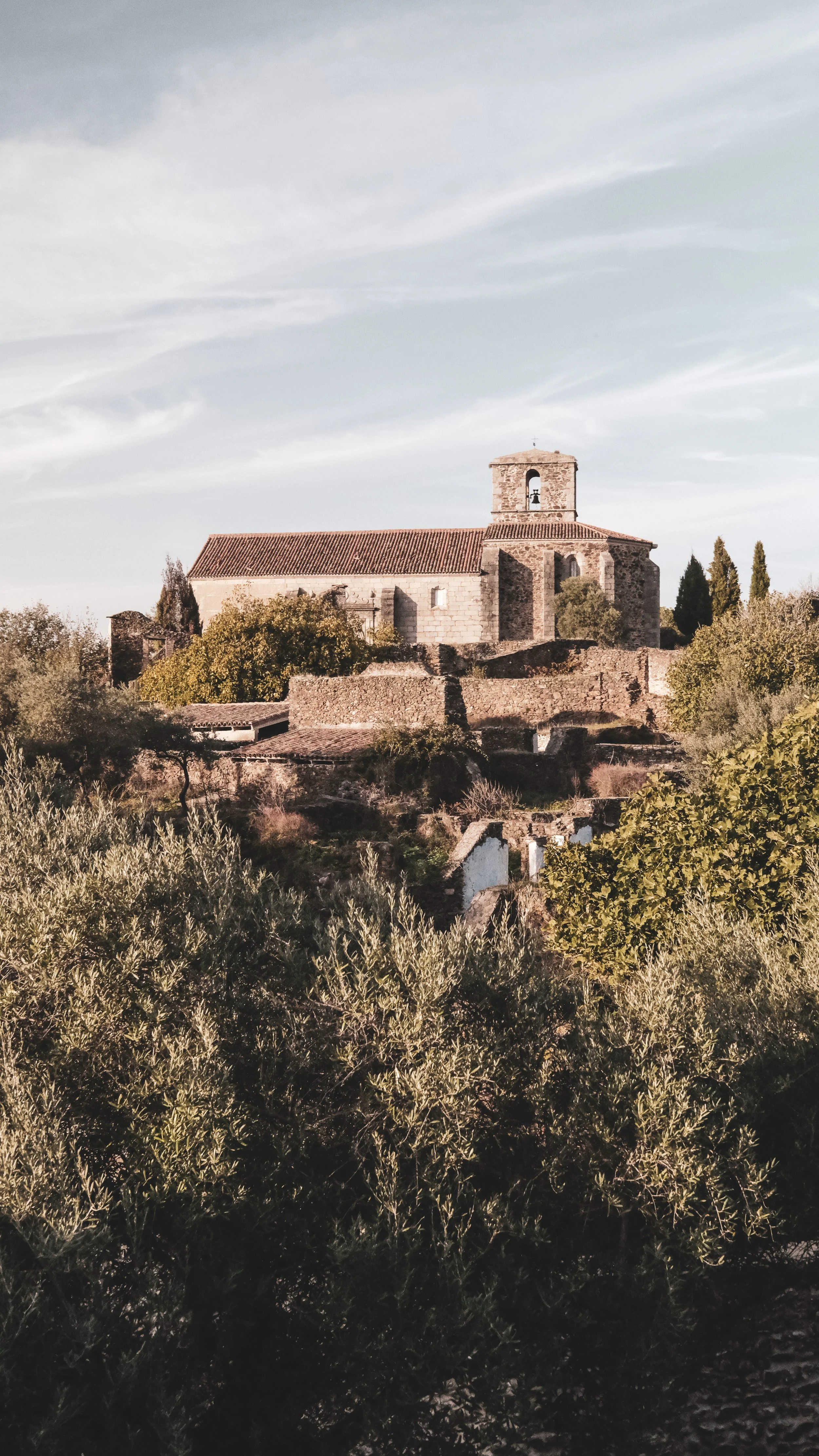 An old stone church on a hill, surrounded by trees and bushes, under a partly cloudy sky.