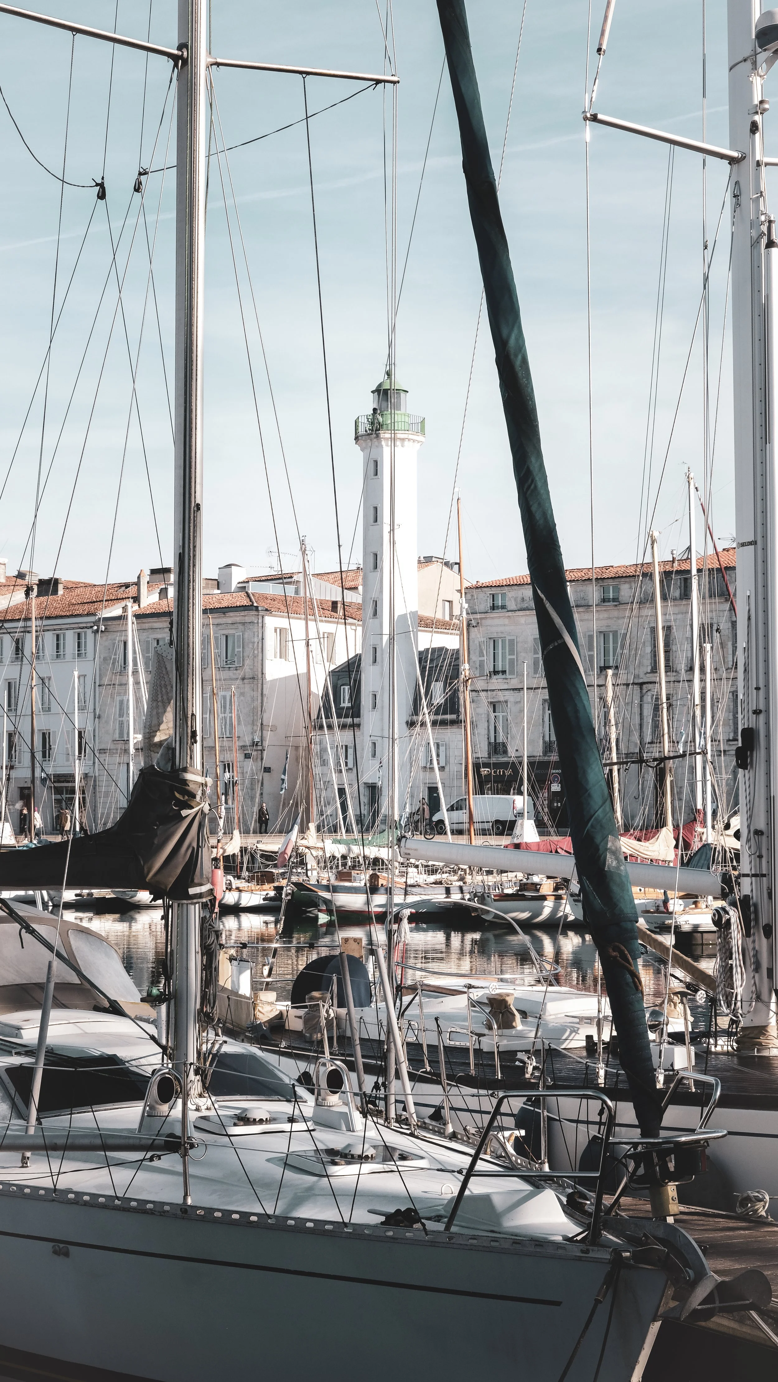 A marina filled with sailboats docked in calm water, with a lighthouse and buildings in the background.