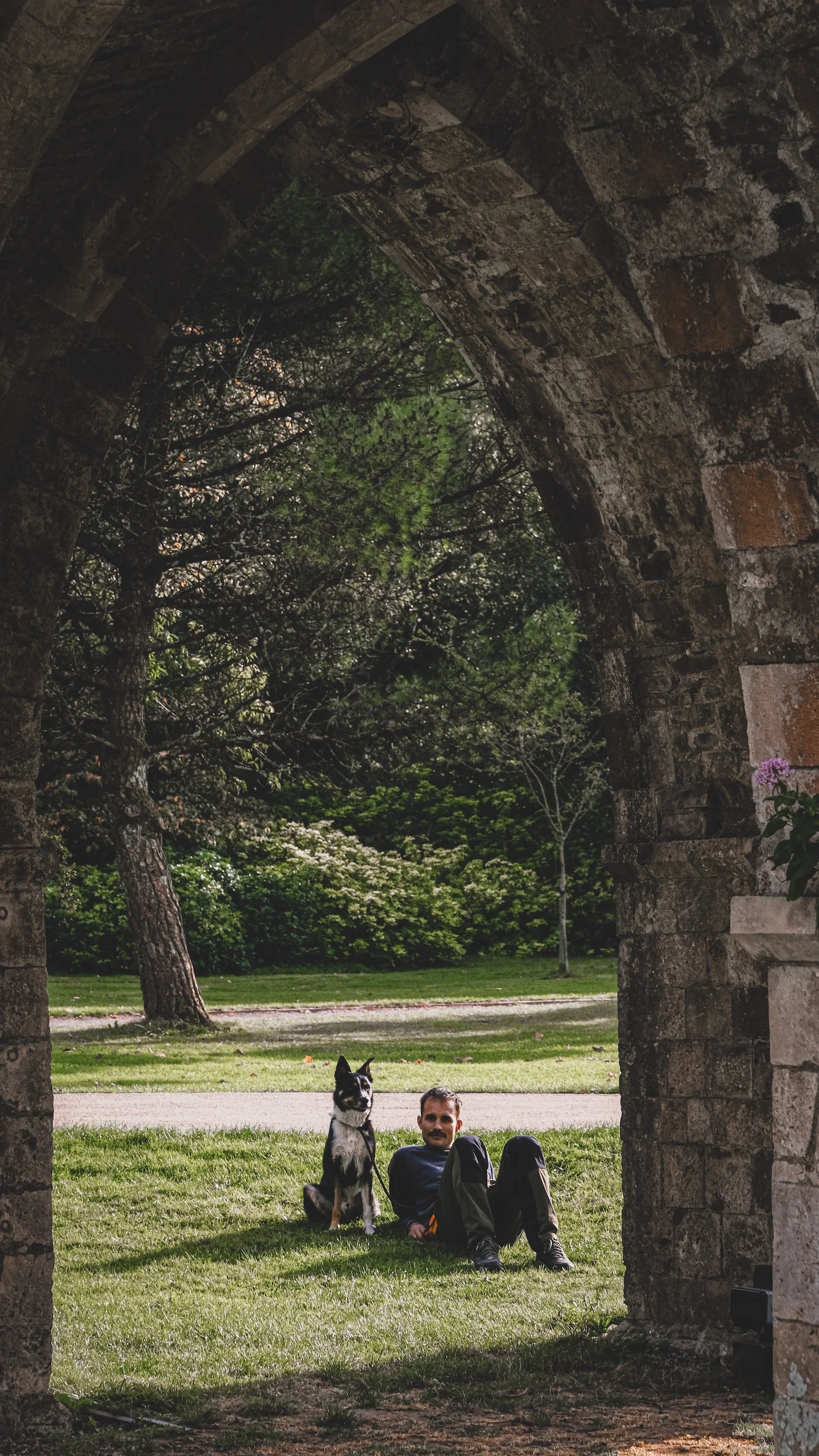 A man and a black and white dog sitting on the grass under a stone archway with trees and a pathway in the background.