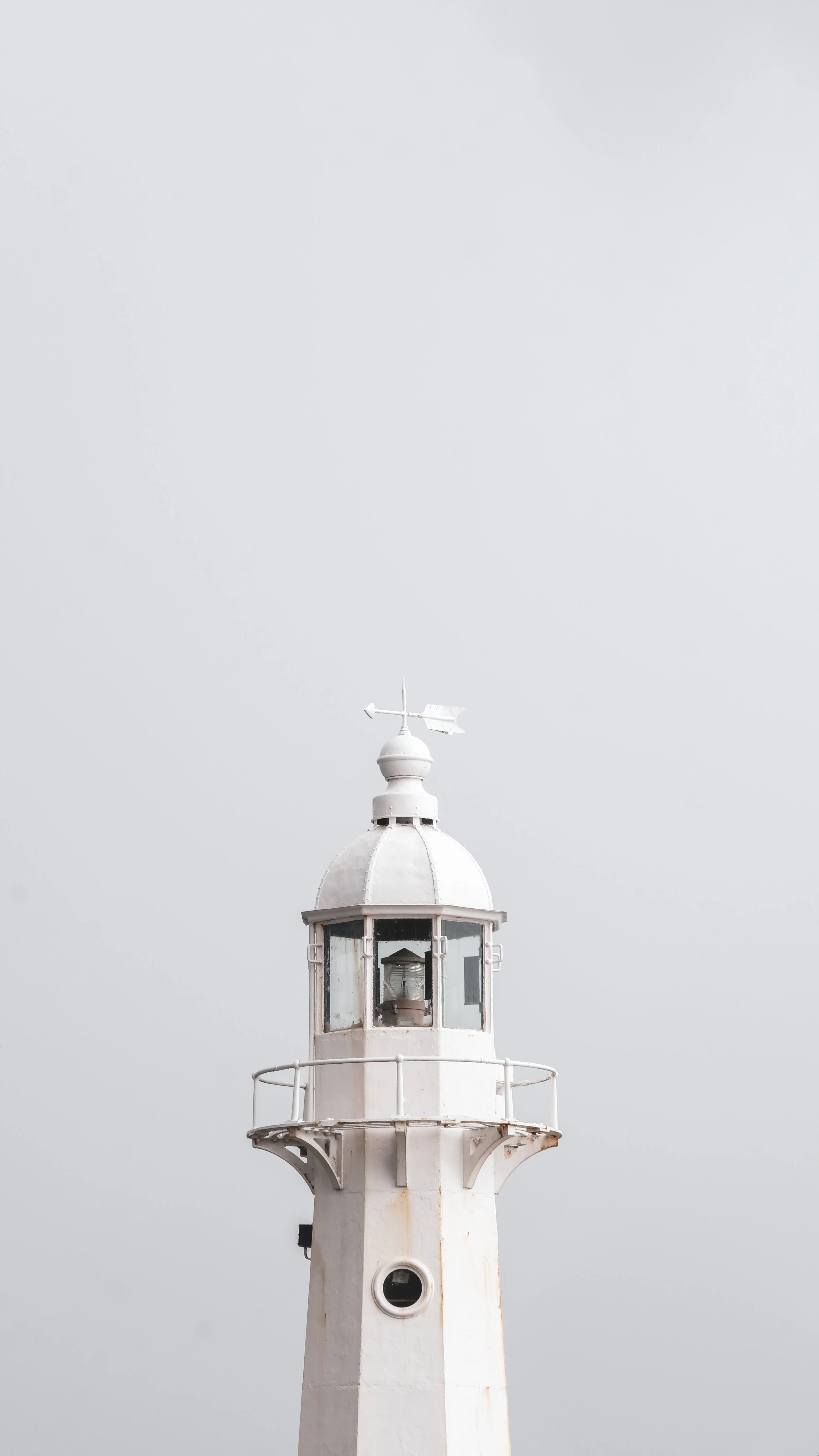 A white lighthouse with a rounded top and a railing around the lantern room, standing against a gray sky.