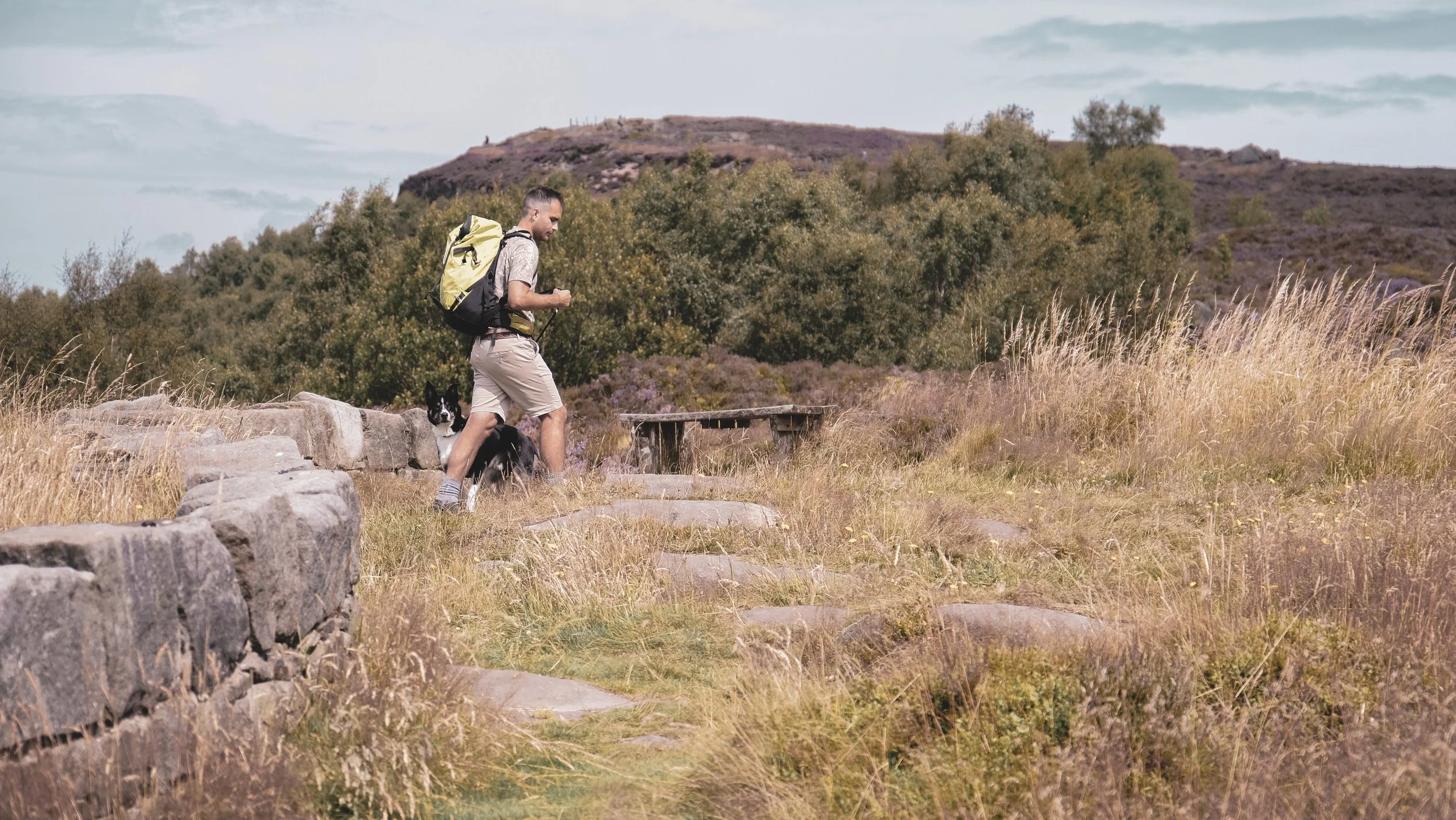 A man hiking with a backpack accompanied by a black and white dog on a scenic trail with grassy and rocky terrain, trees, and rolling hills in the background.
