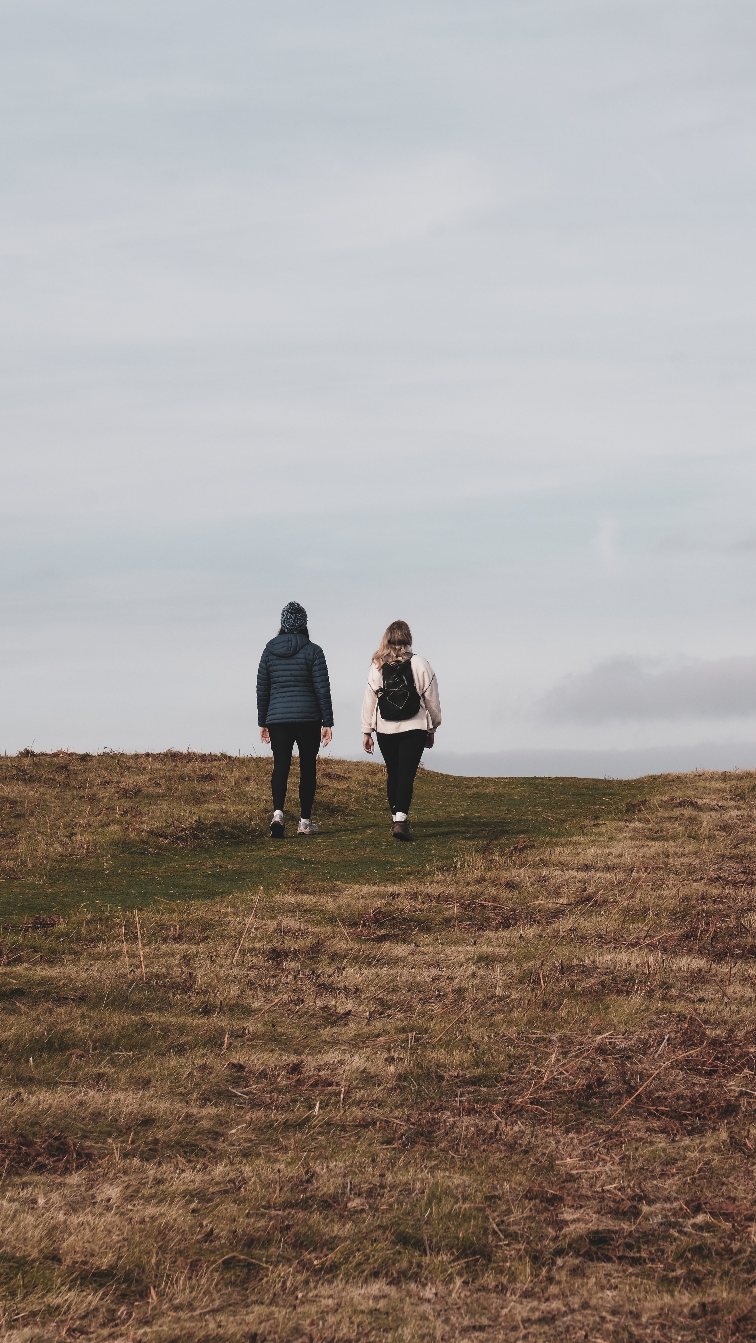 Two women walking through a grassy field towards the horizon under a cloudy sky.
