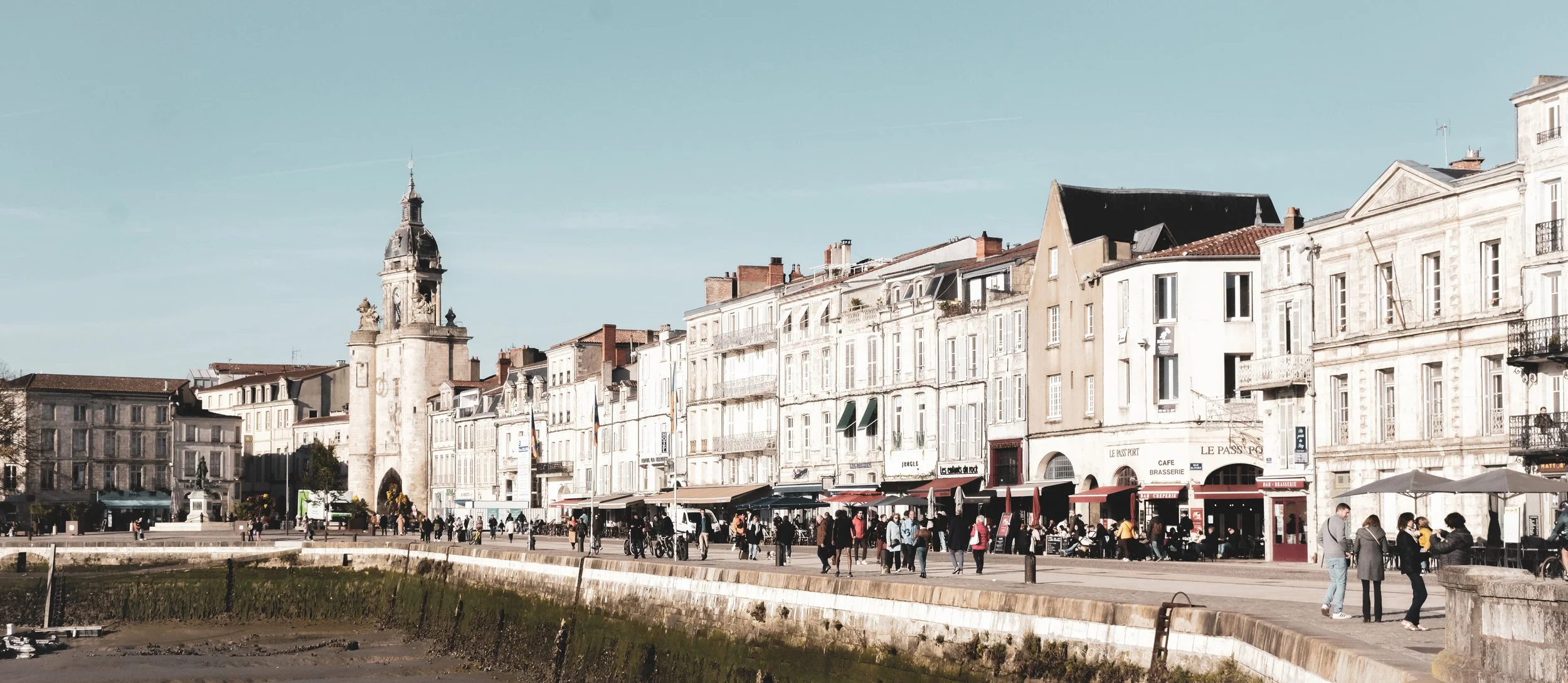 Image of a European city waterfront with historic multi-story buildings, cafes, and people walking along the promenade under a clear blue sky.
