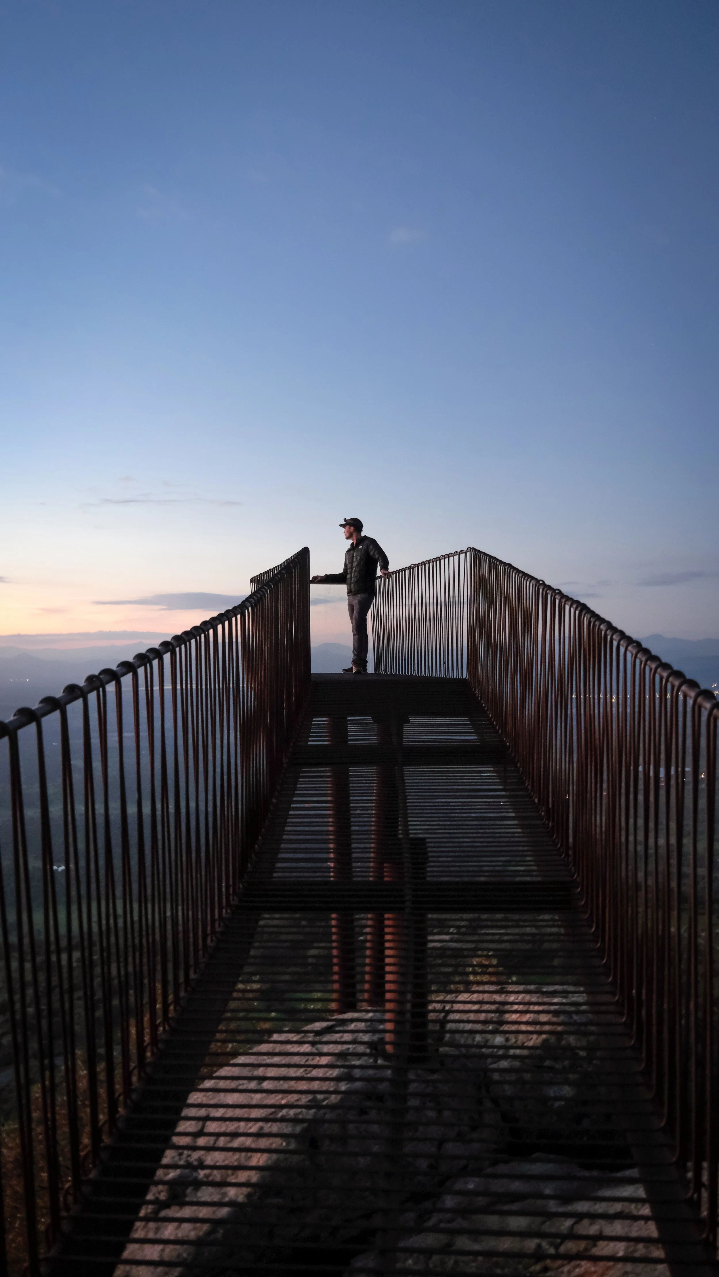A person standing on a metal observation platform at sunset with a mountain view in the background.
