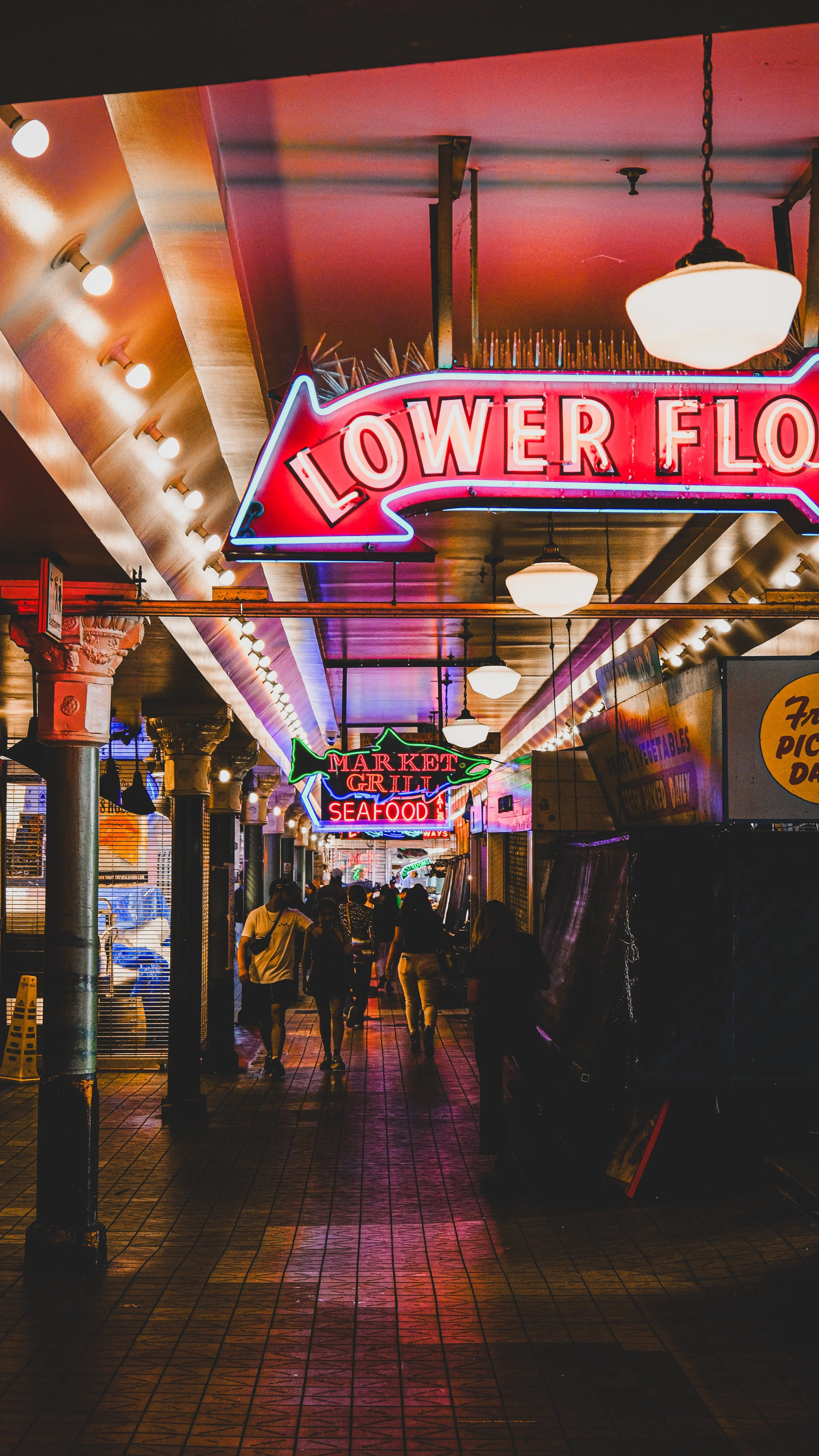 Night market with neon signs for 'Lower Floor' and 'Market Grill Seafood', walkways lined with people, lit by hanging lights, stalls on the sides.