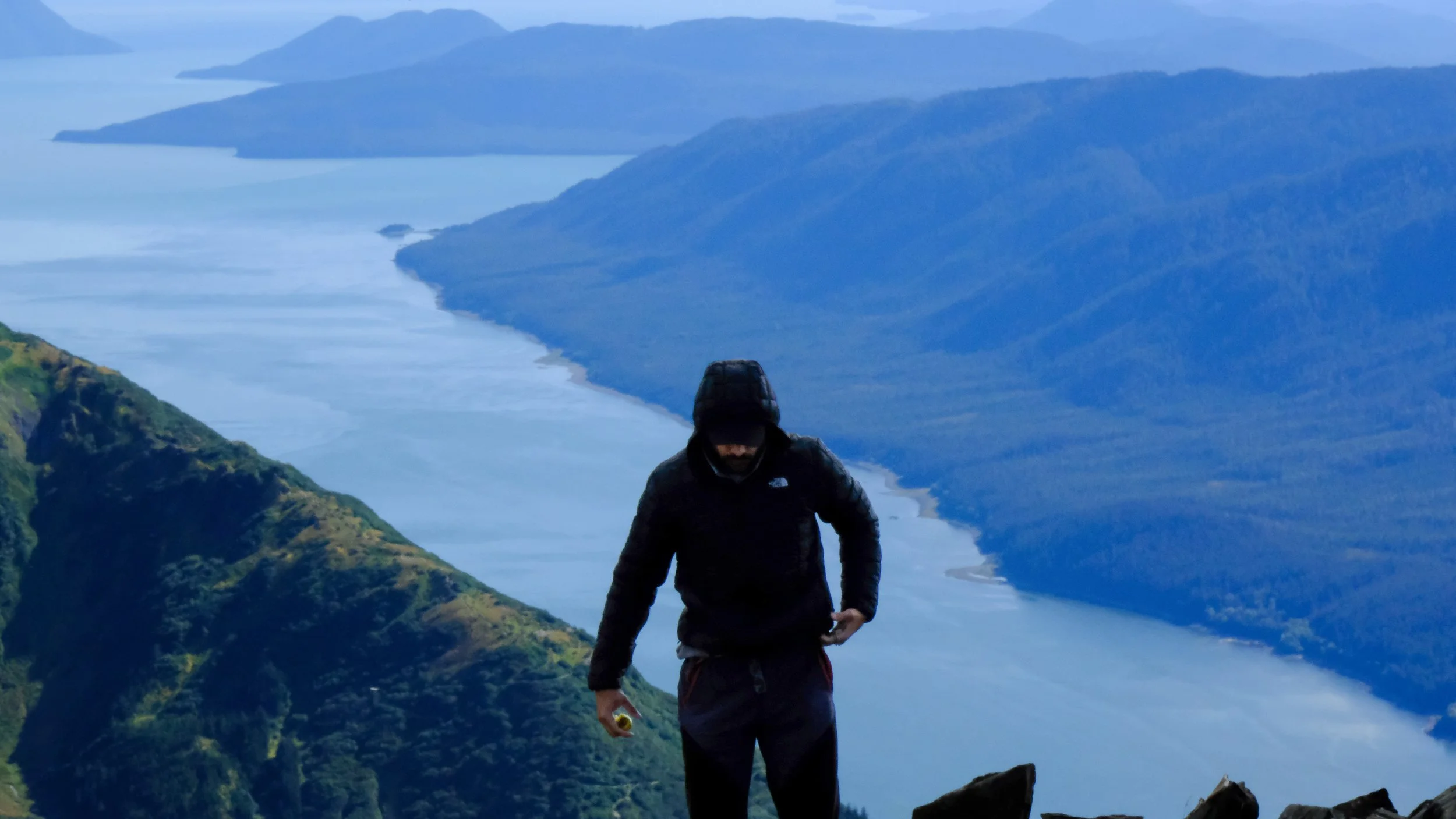 Hiker dressed in black jacket and hood standing on mountain trail overlooking river and forested hills in the distance.