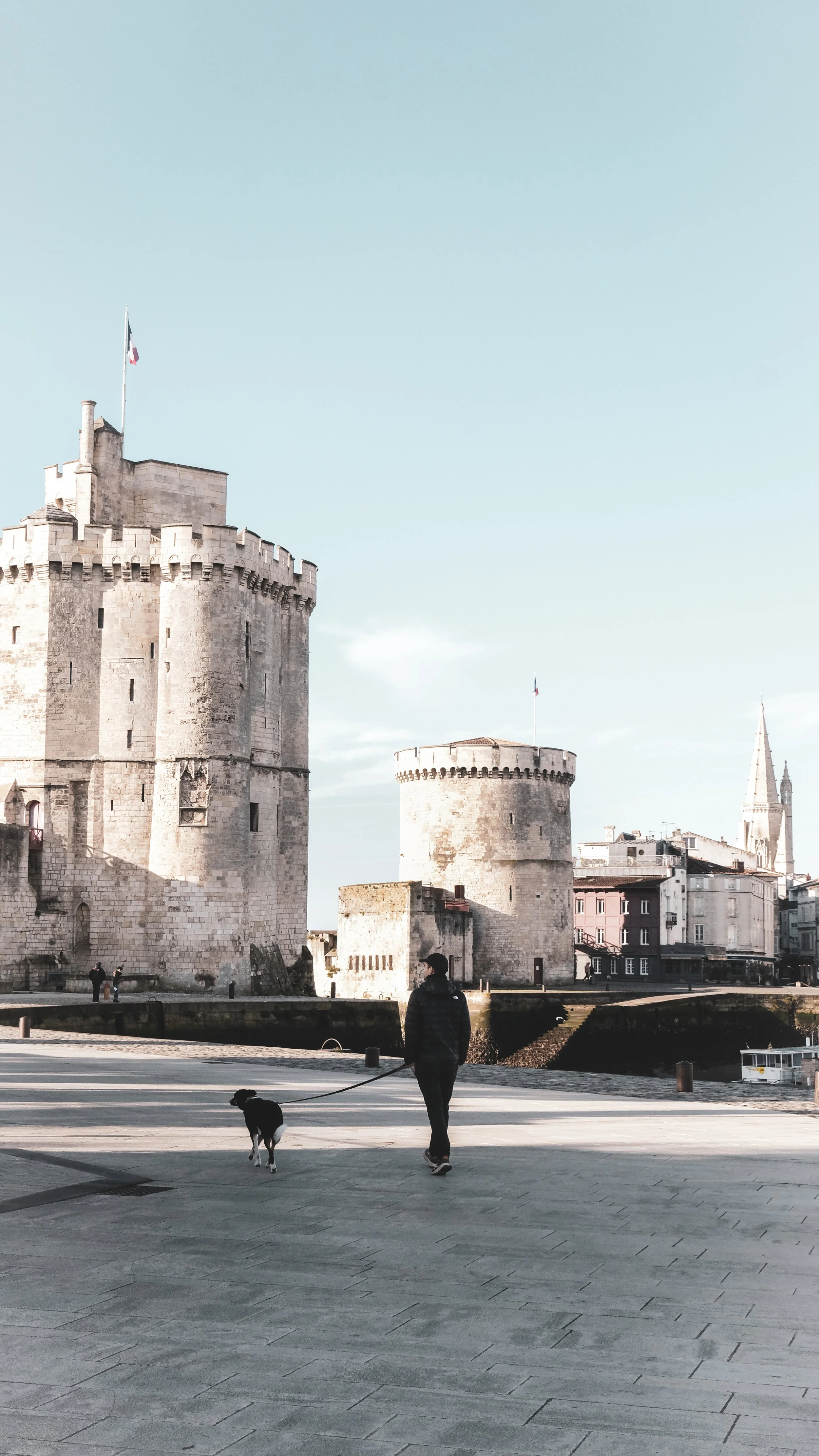 A person walking a dog along a paved promenade near historic stone castles with a tall church steeple in the background on a clear day.