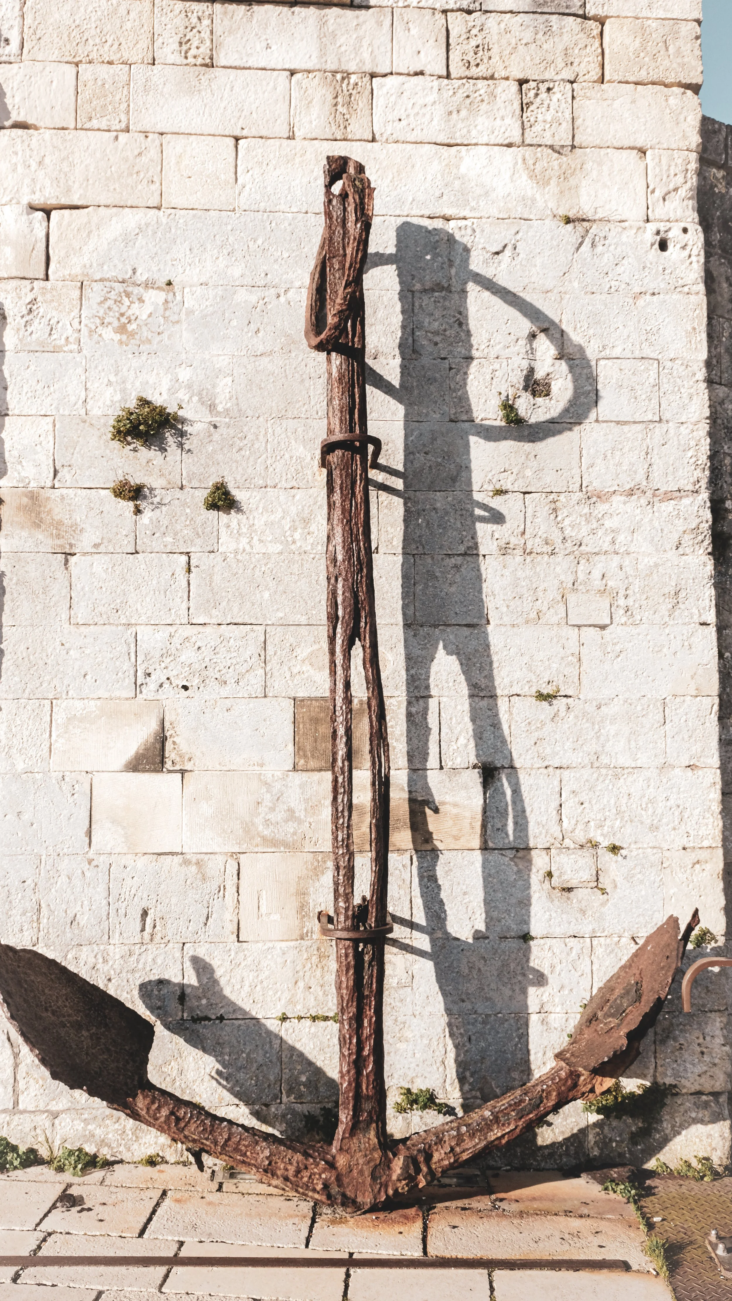 Old rusted anchor leaning against a stone wall with its shadow cast on the wall.