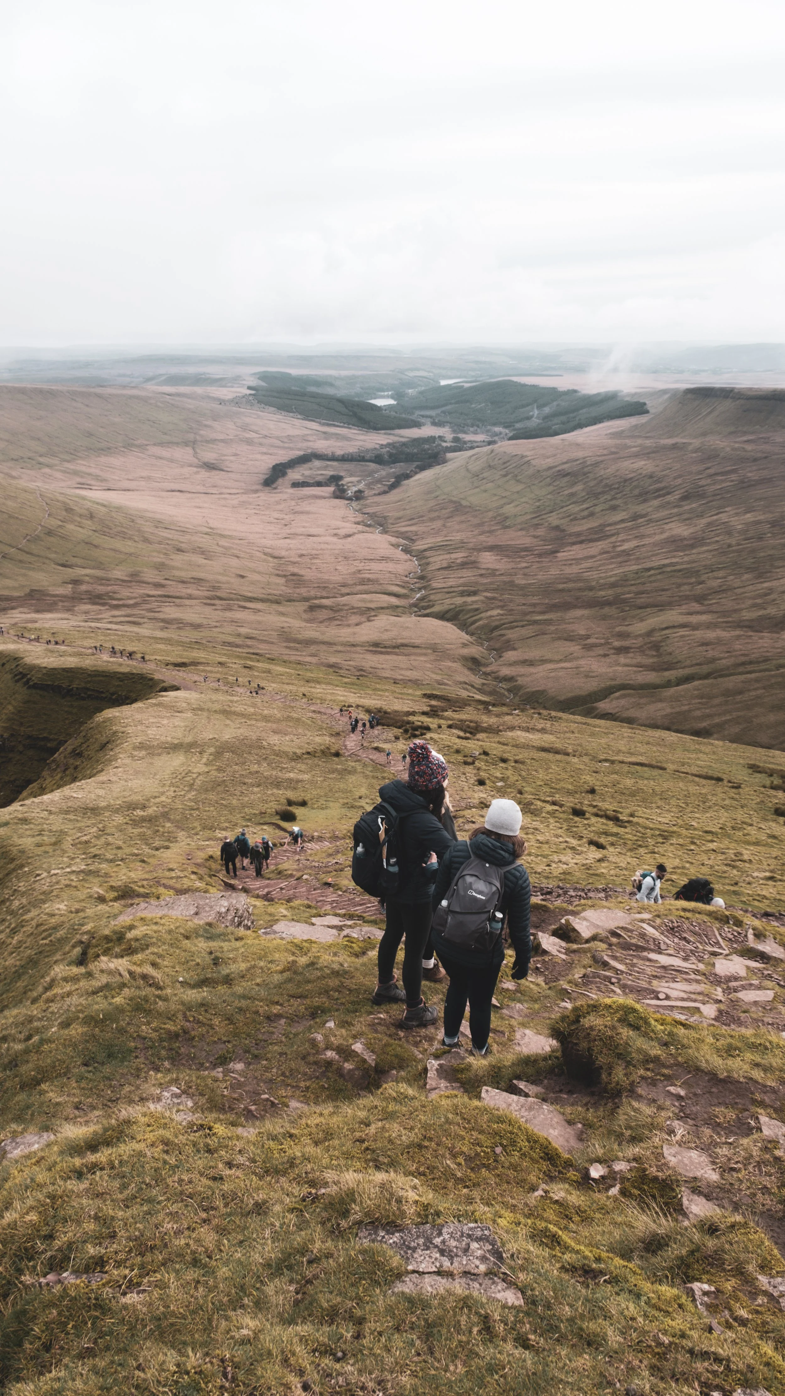 Hikers walking down a trail in a hilly, grassy landscape with a river in the distance and cloudy sky.