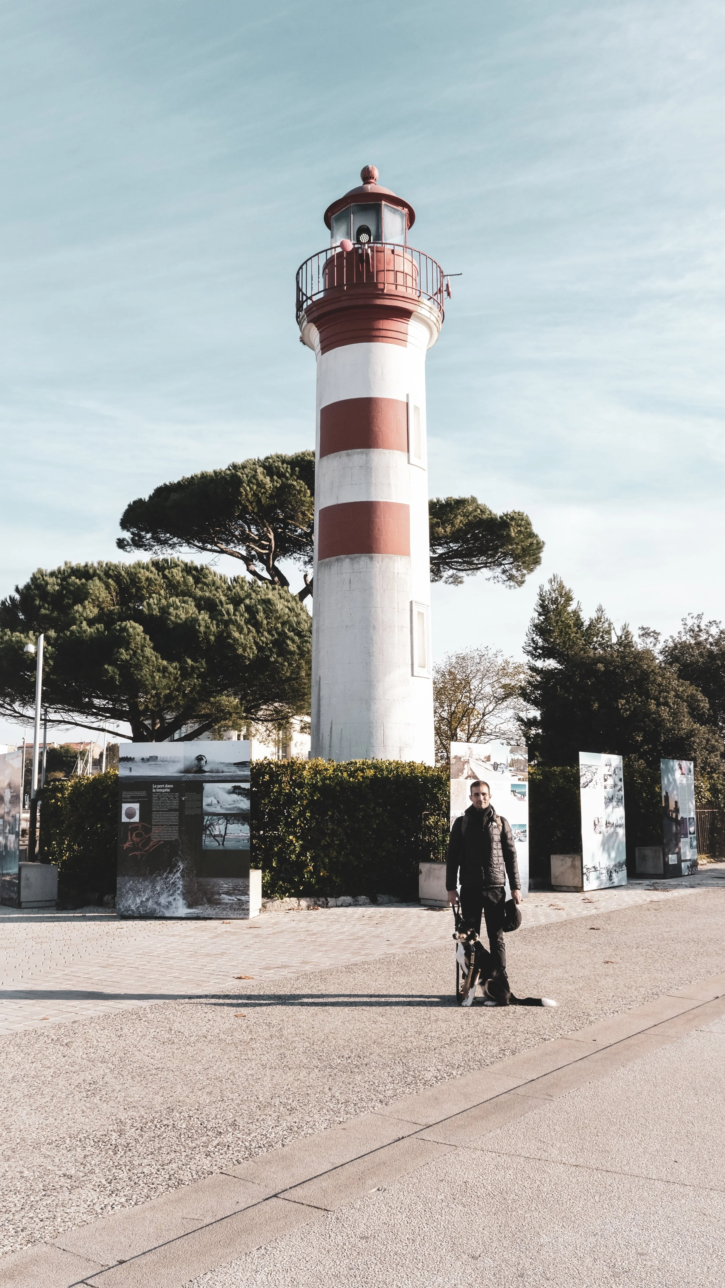A man standing with a dog in front of a tall red and white striped lighthouse with trees and informational panels in the background.