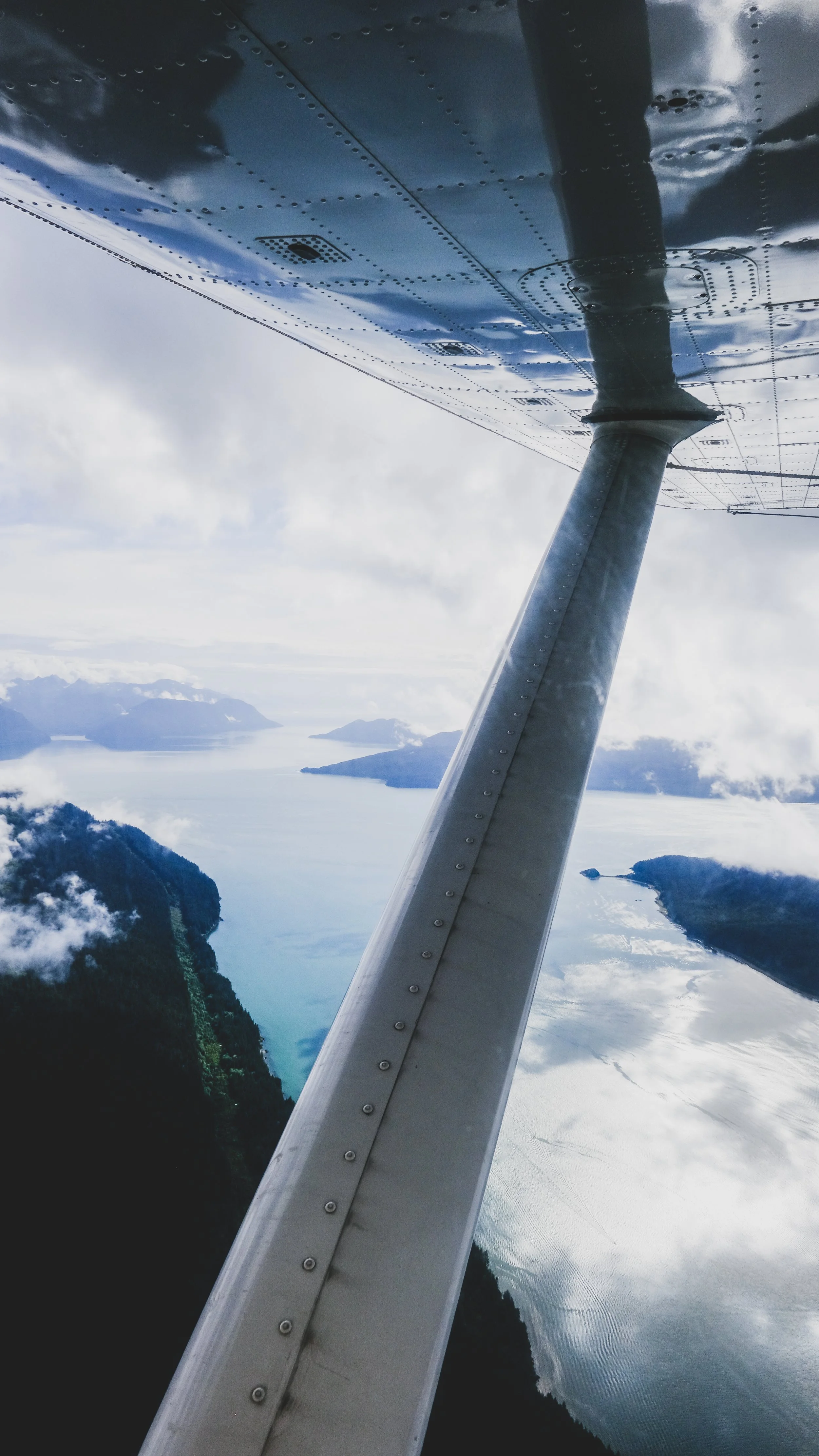 View from a small plane window over a body of water with islands, mountains, and clouds in the sky.