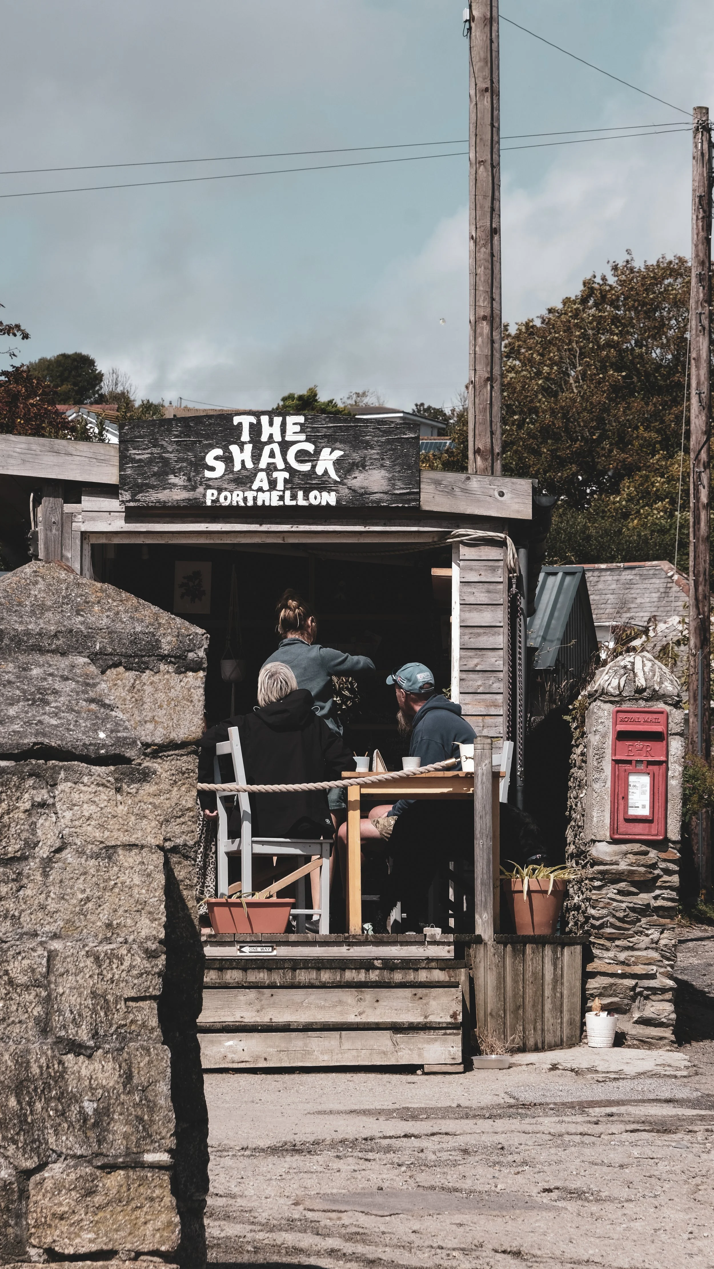 People dining and chatting at an outdoor wooden restaurant called The Shack at Portellon. The restaurant has a rustic appearance with a weathered sign and is surrounded by a stone wall and wooden posts.
