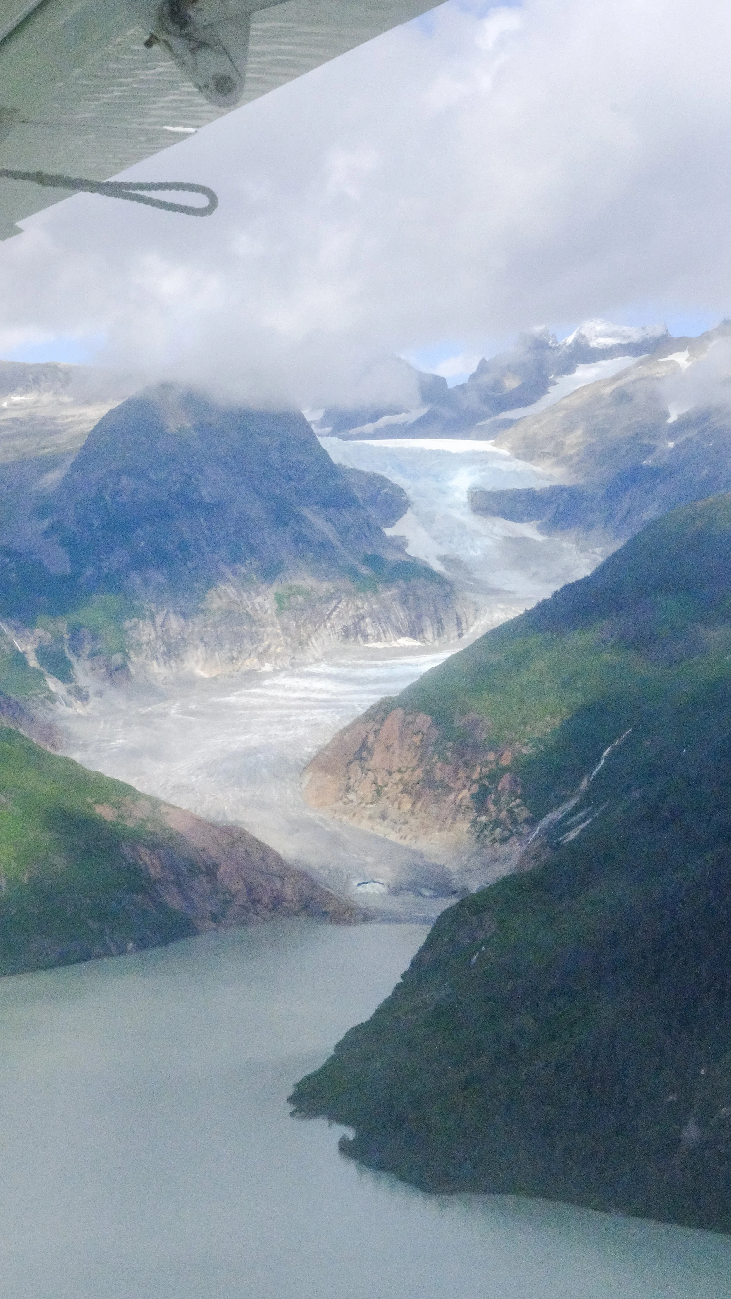 Aerial view of a glacier surrounded by green mountains and a lake below, with cloud cover in the sky.