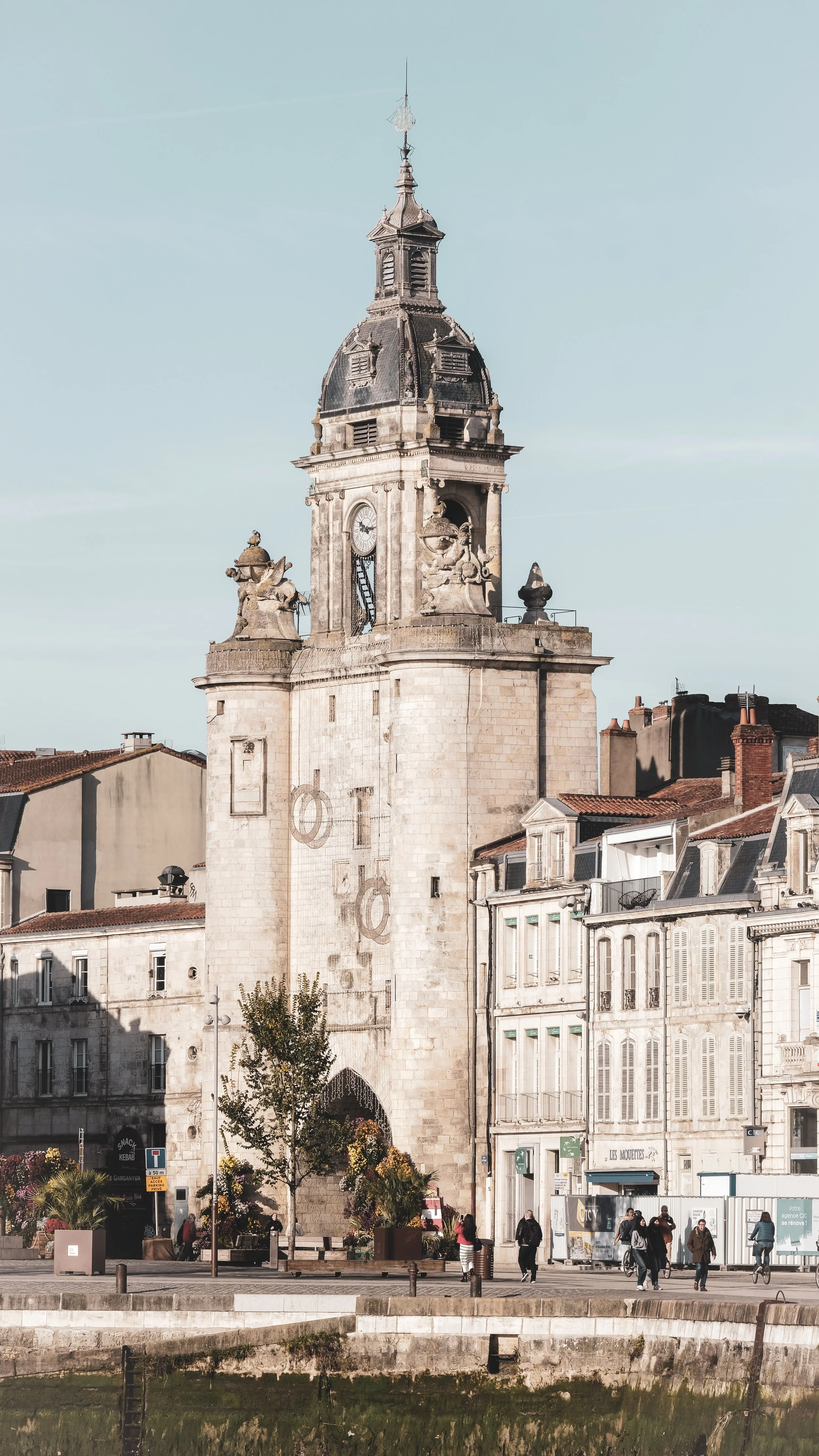 An old stone clock tower on a city street with buildings and pedestrians, under a clear blue sky.