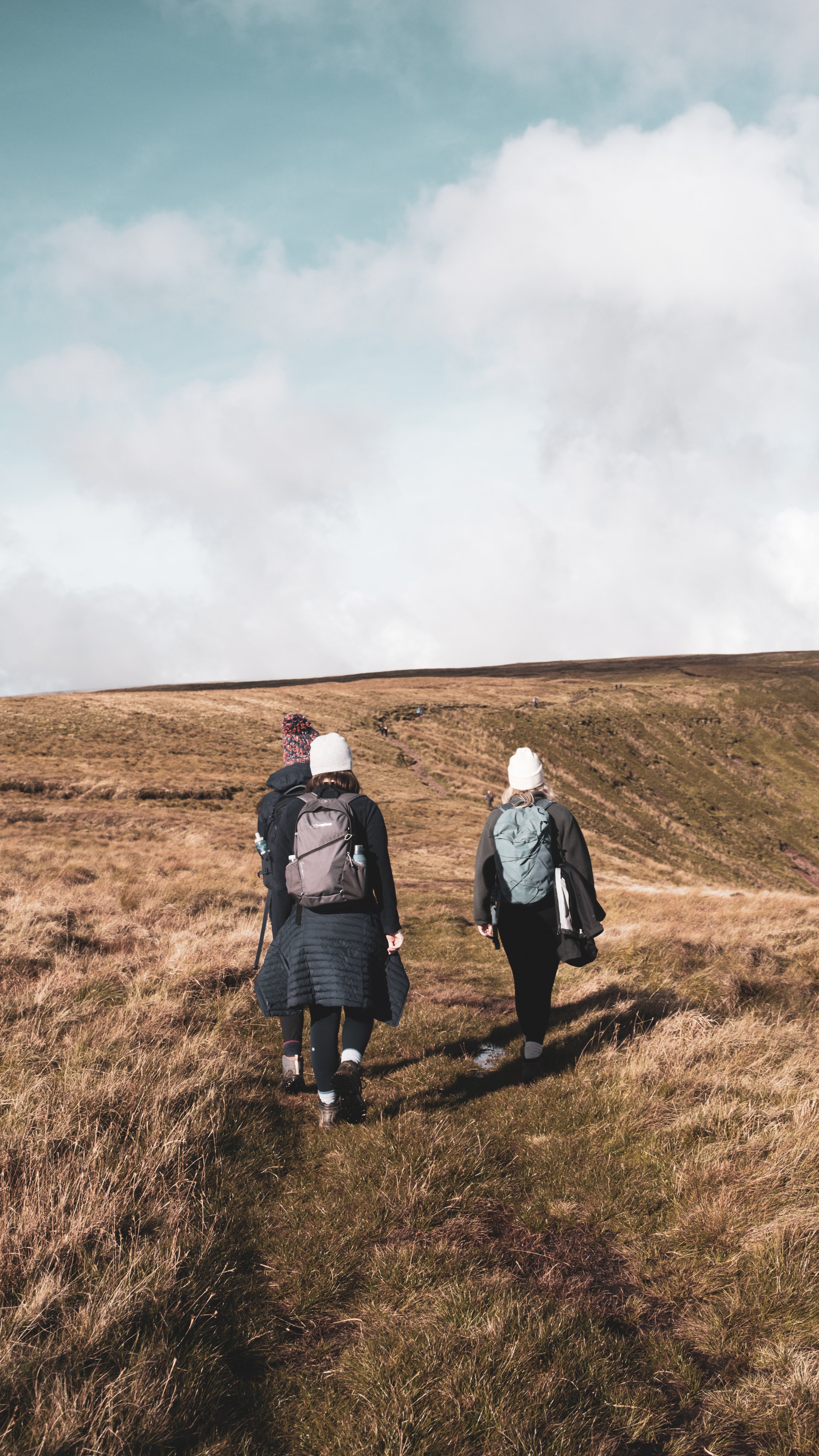 Three hikers with backpacks walking on a trail through a grassy landscape under a partly cloudy sky.