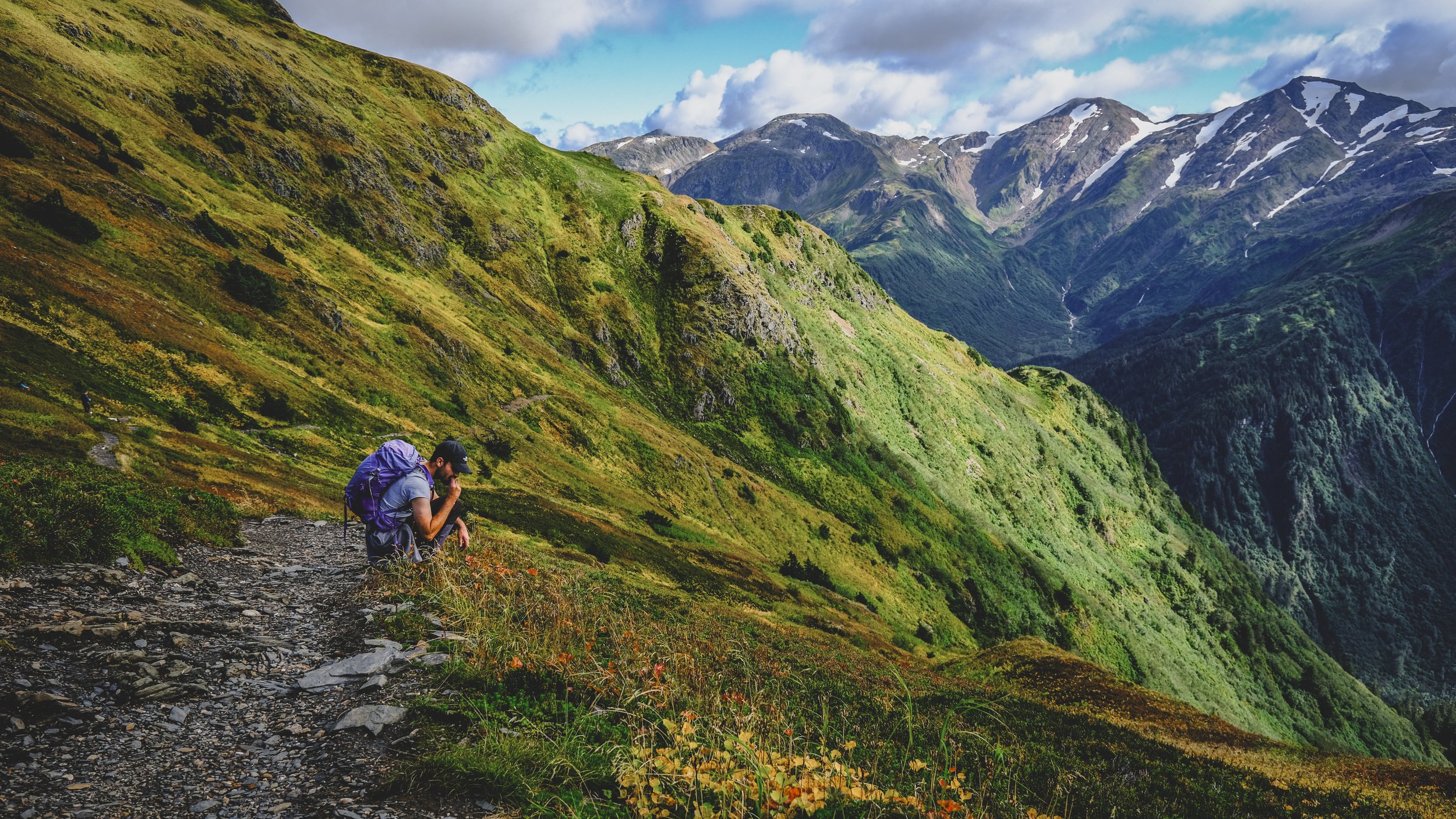 A hiker with a purple backpack kneeling on a rocky mountain trail surrounded by green mountains and a partly cloudy sky.