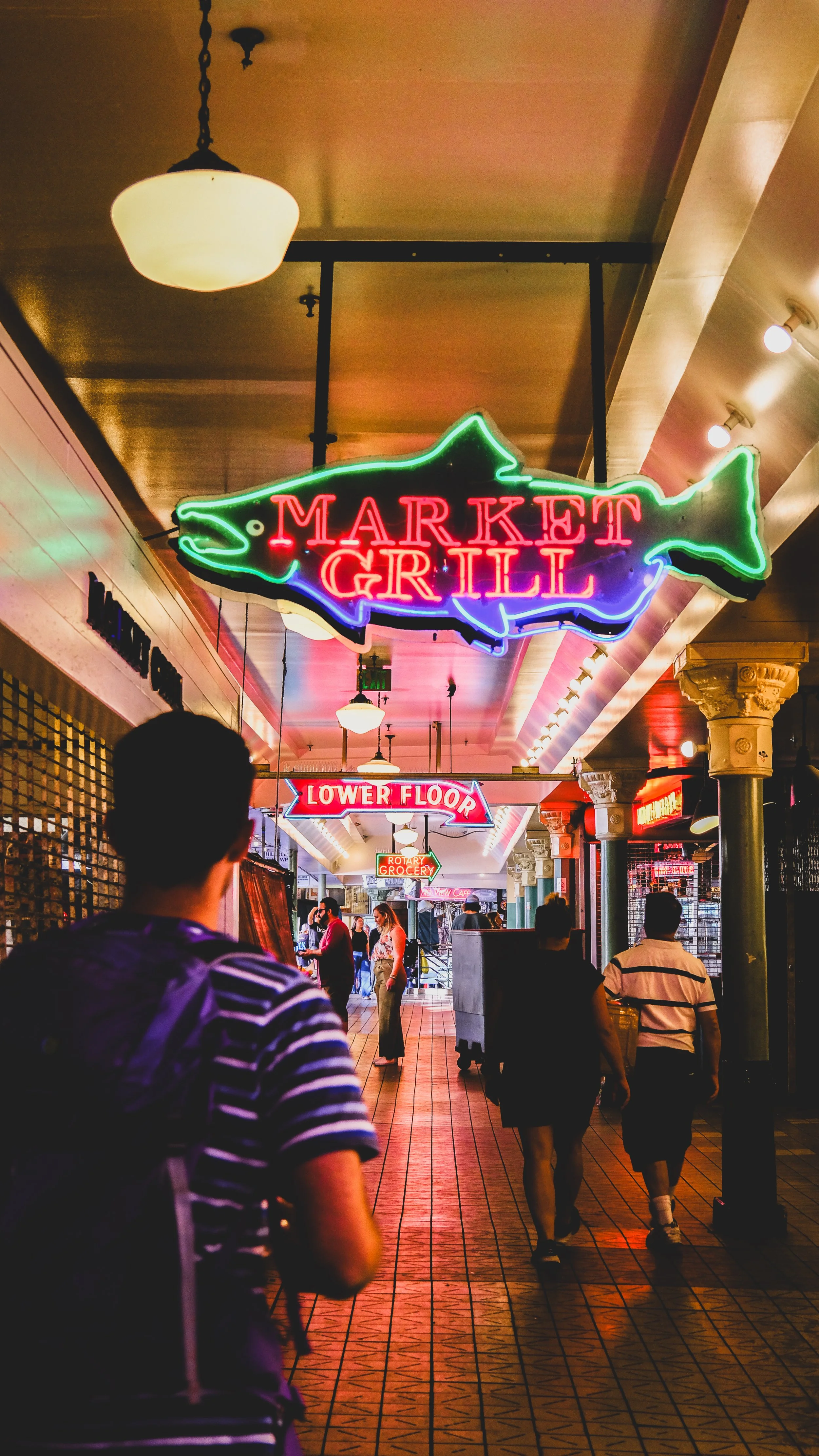 Neon signs hanging in an indoor marketplace or mall corridor, including a fish-shaped sign reading 'Market Grill' and another for the 'Lower Floor' with people walking underneath.