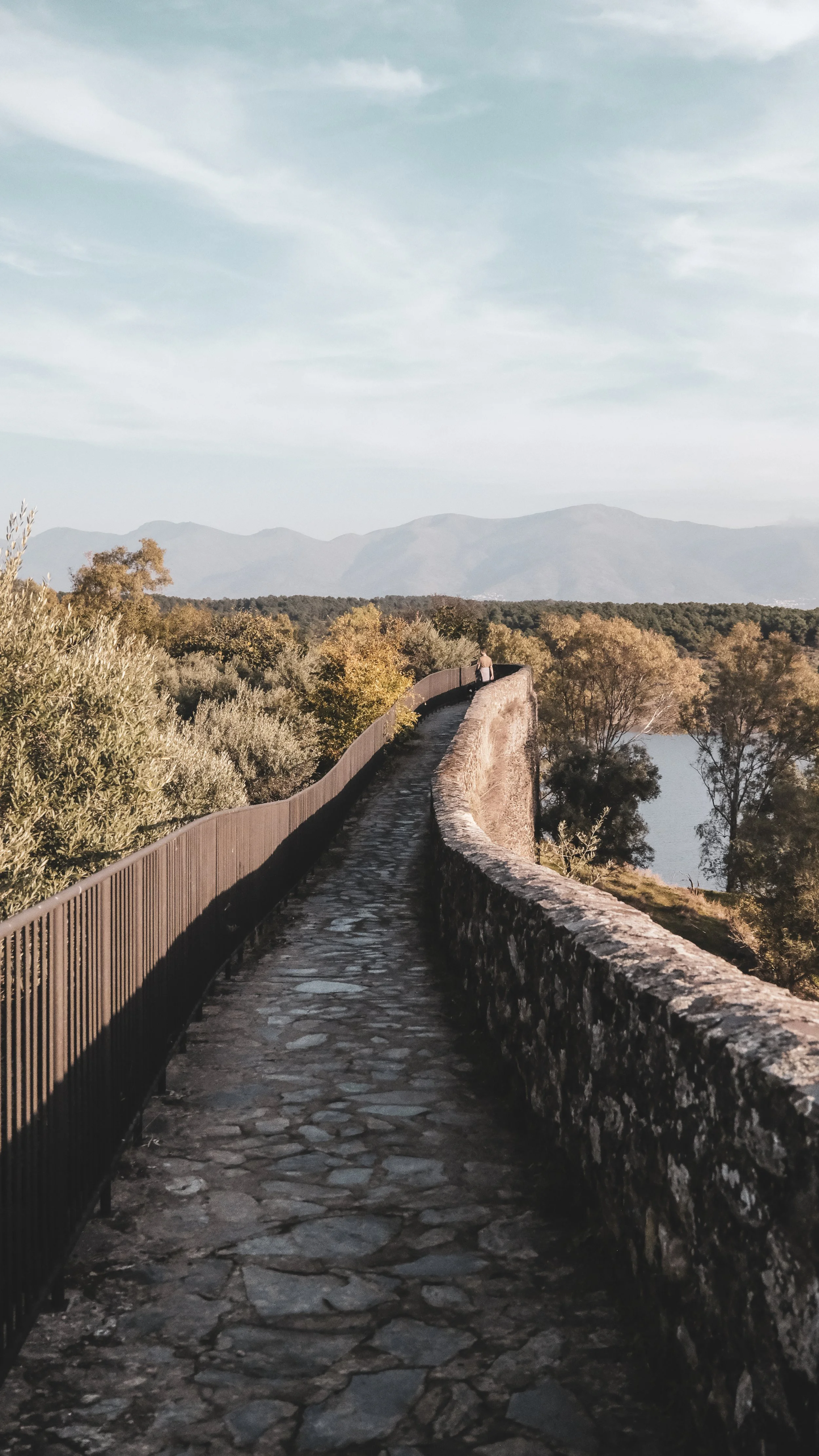 A cobblestone pathway with stone and metal railings curving along a hillside overlooking a body of water, with trees and mountains in the distance under a partly cloudy sky.