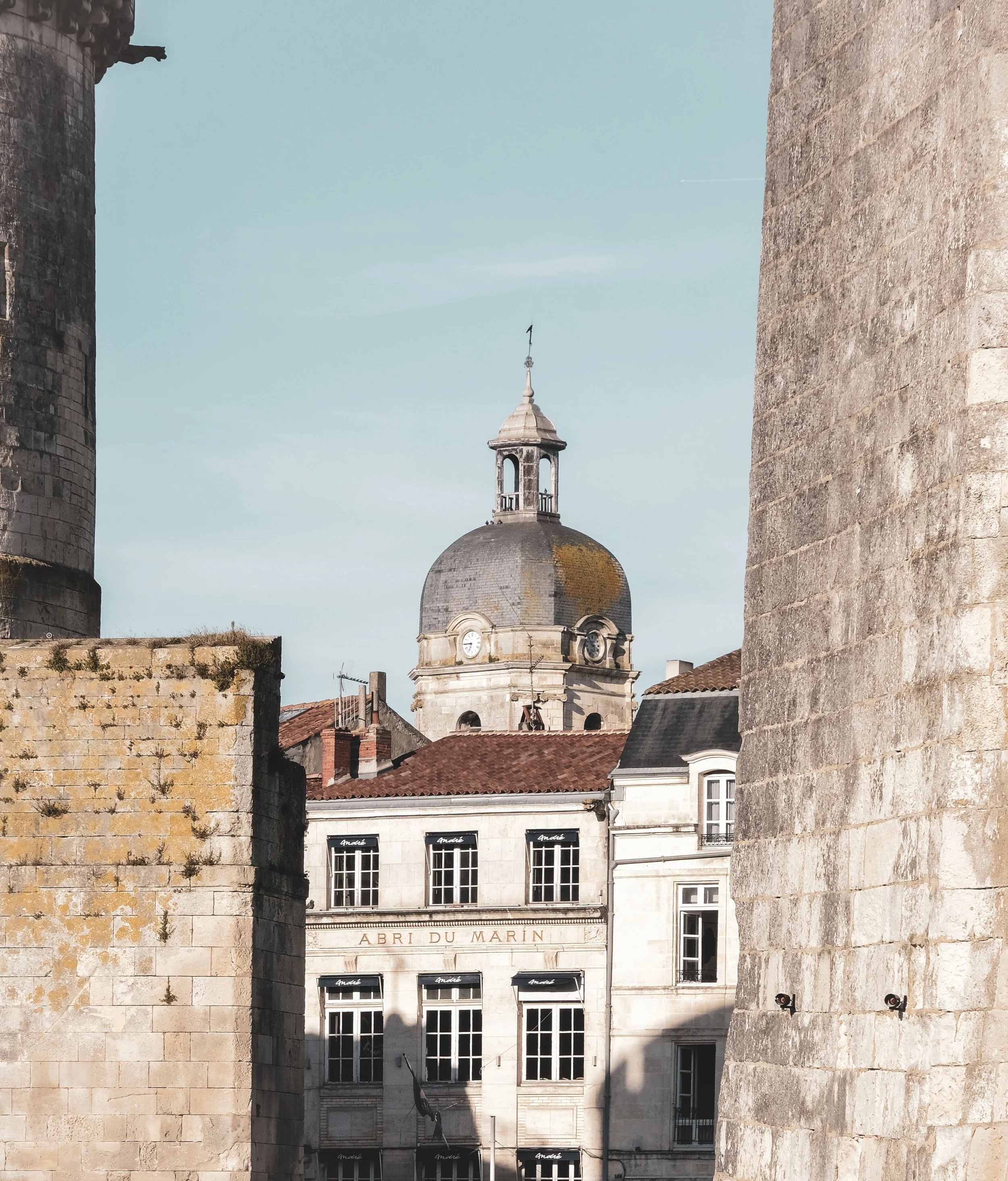View of a historic building with a domed clock tower, framed by ancient stone walls. The building has a sign that reads 'ABRI DU MARIN' and features multiple windows and a red-tiled roof.