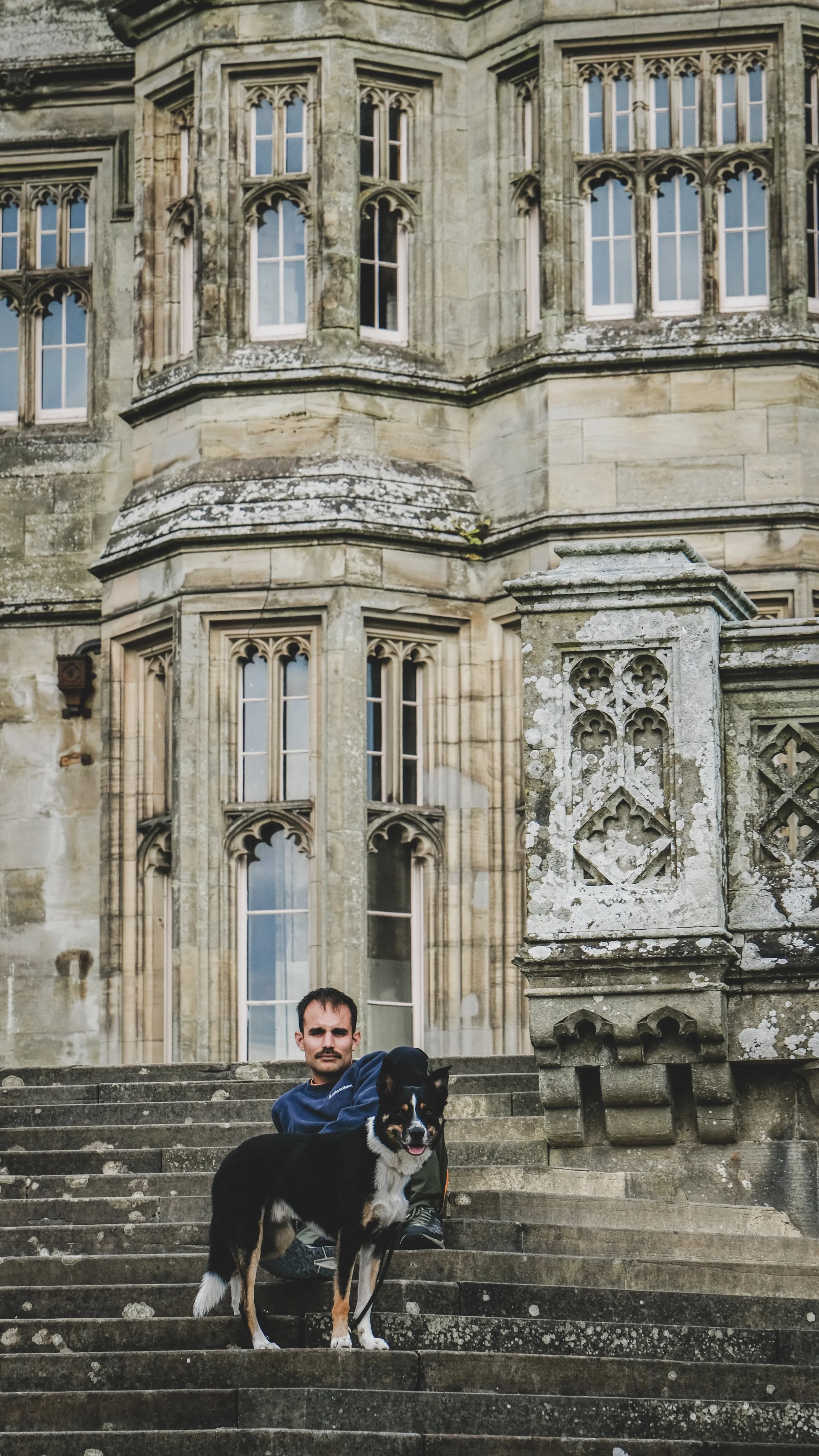 A man with dark hair and a mustache sitting on stone stairs with a black and white dog in front of a historic stone building with detailed gothic windows and architecture.