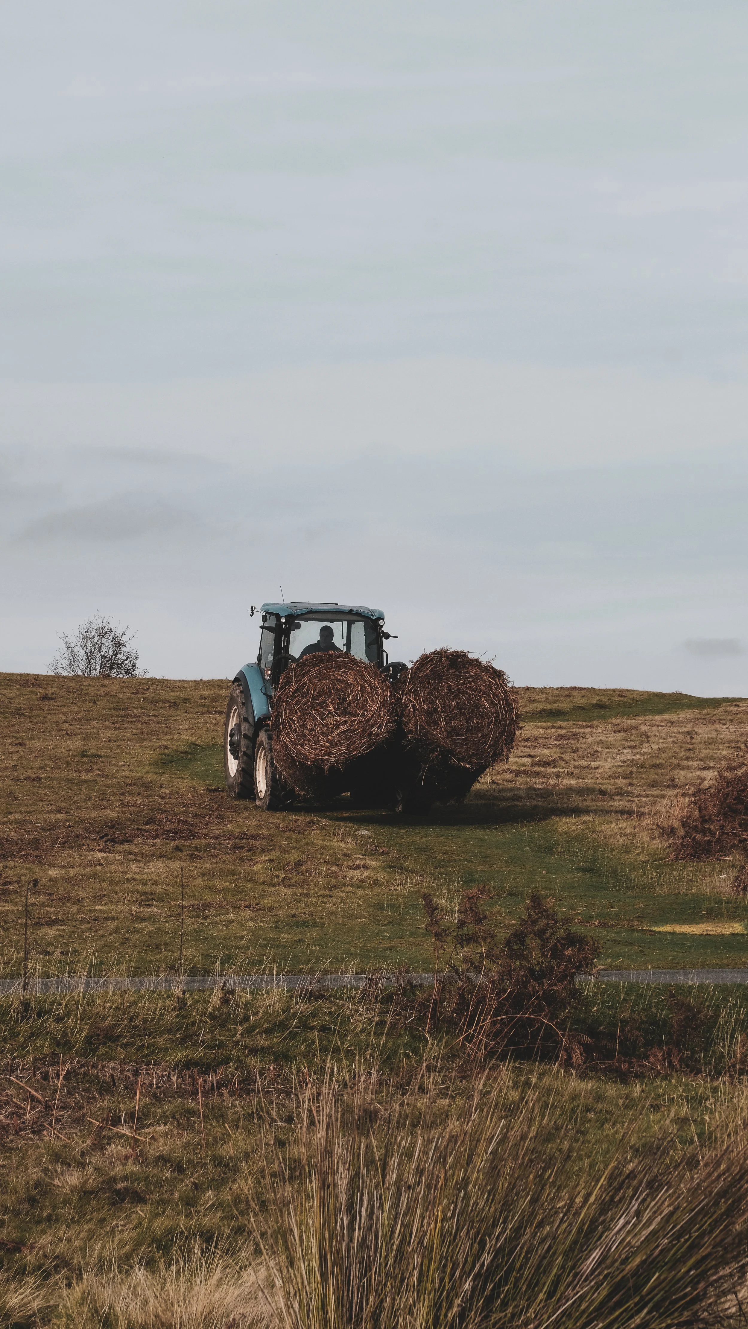 A tractor carrying two large rolled hay bales on a grassy hill under a cloudy sky.