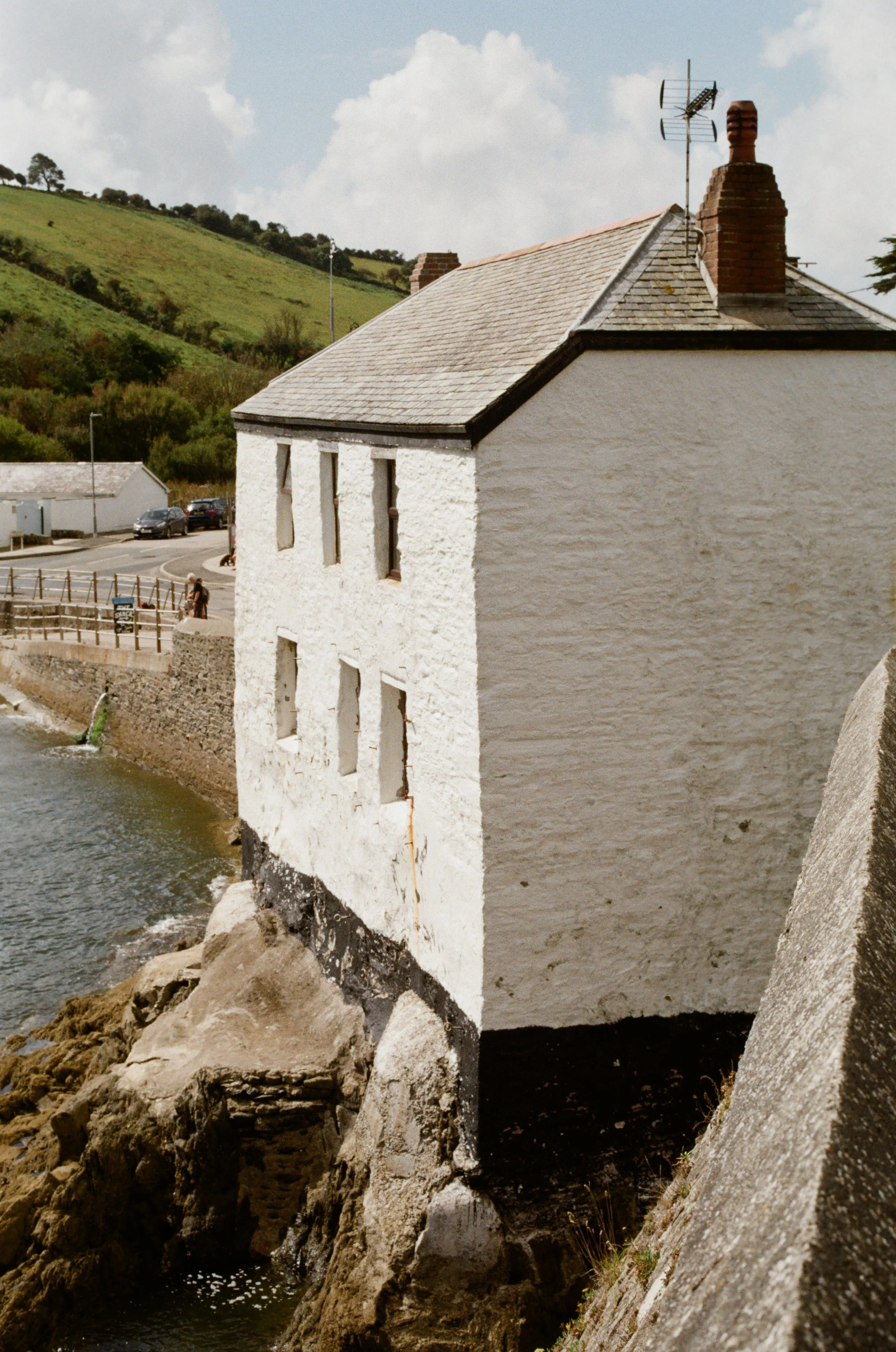 A white, old building partially overhanging a rocky shoreline near the water, with a slate roof and small windows, in a coastal village with hills and a road in the background.