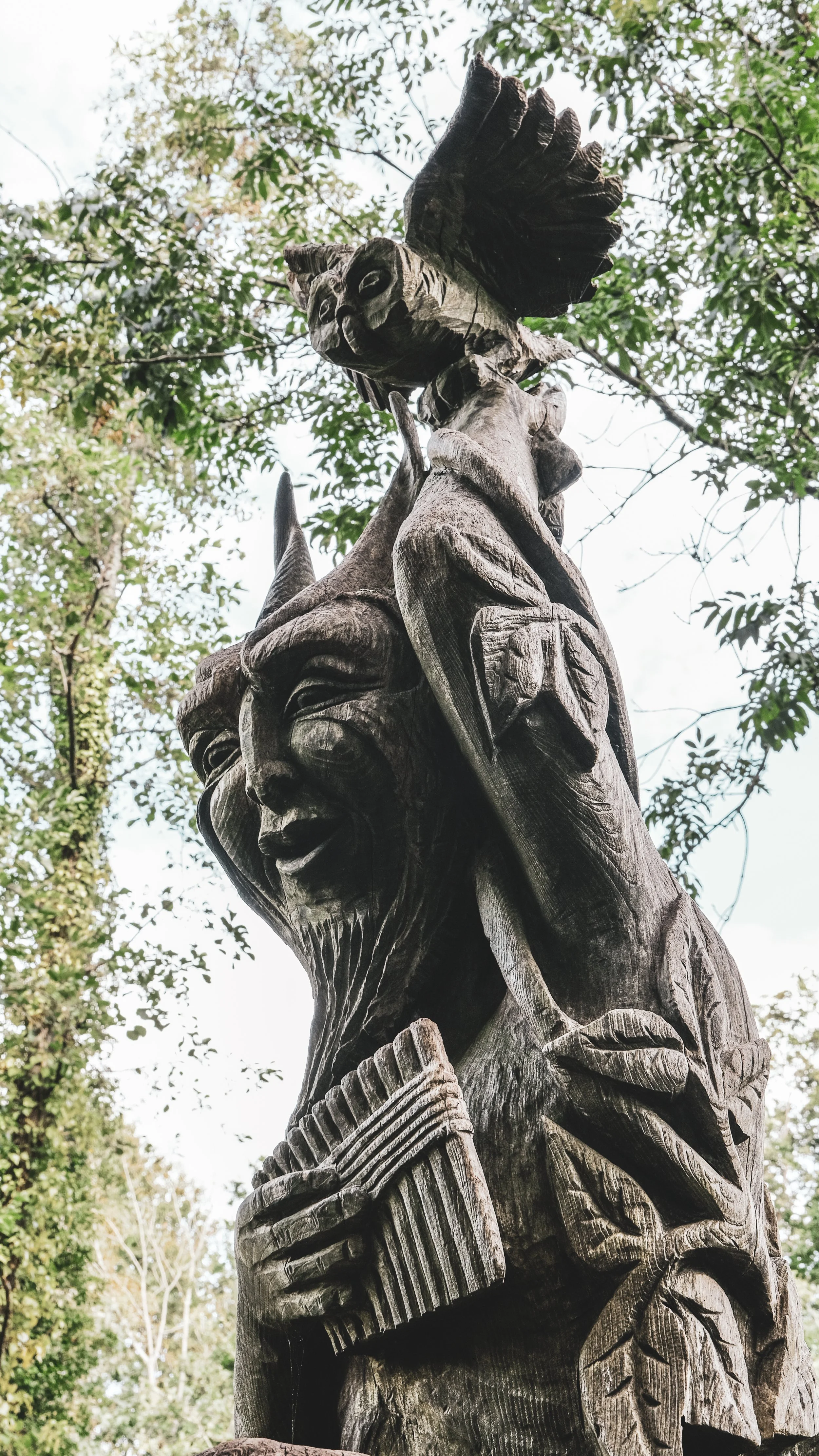 A tall wooden sculpture with carved faces and characters, with detailed features and leaves carved into it, set outdoors among trees.