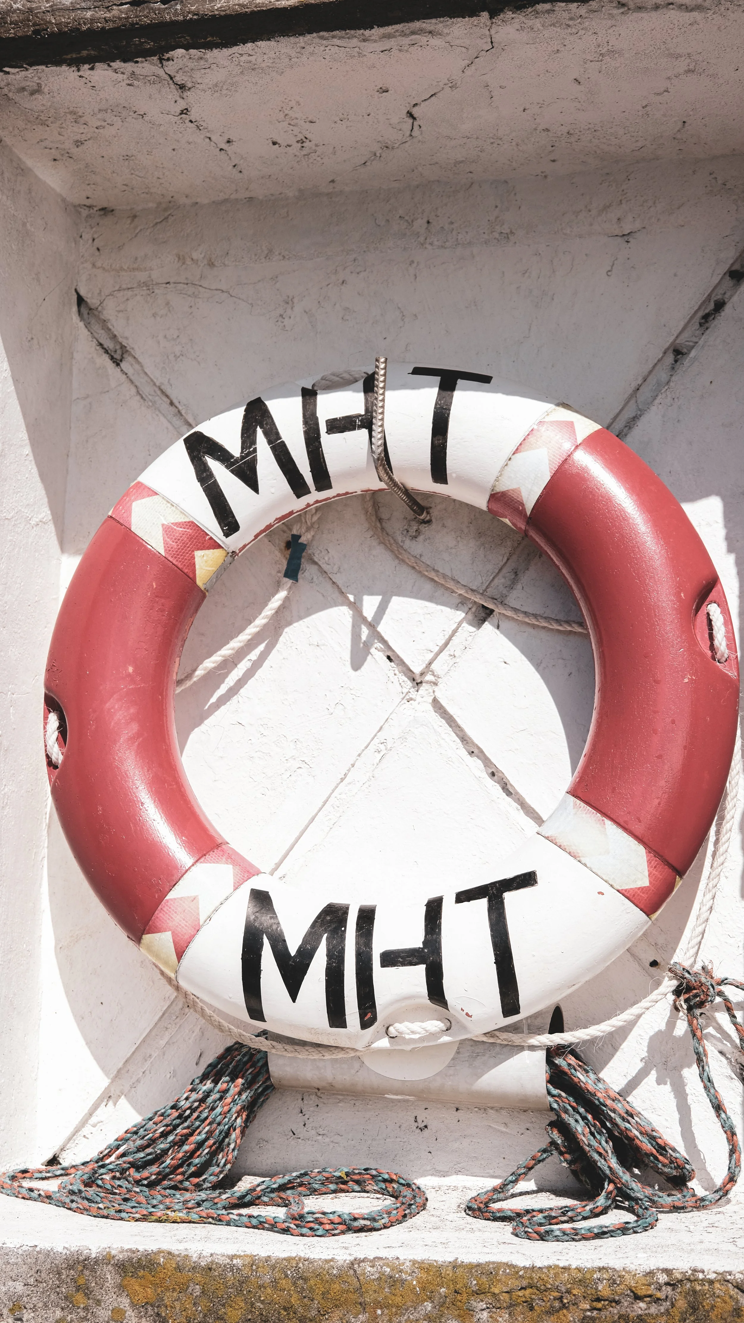 A white lifebuoy with red sections, labeled 'MHT' in black letters, mounted on a white wall with ropes attached.