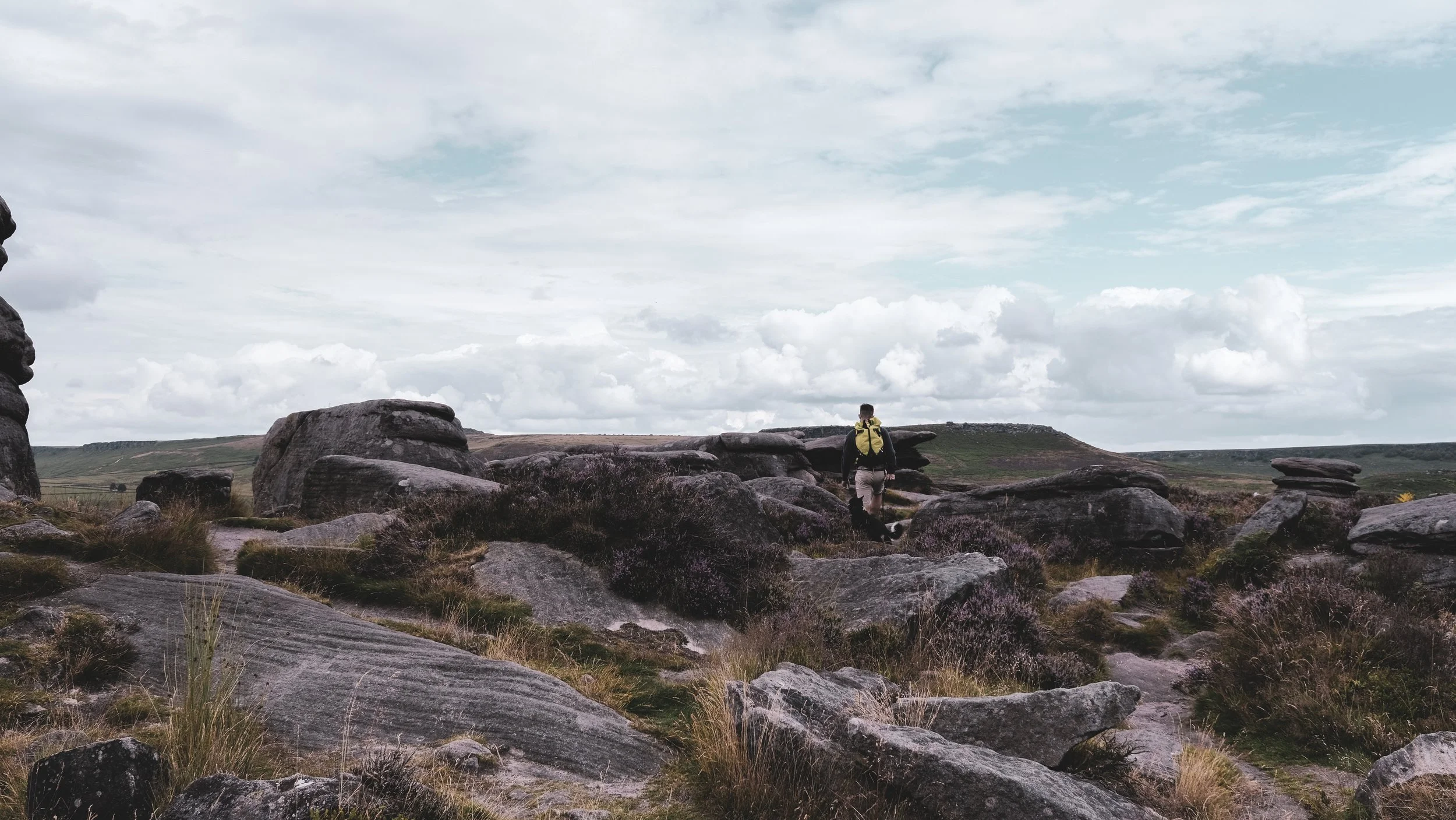 A person with a yellow backpack walking among large rocks and boulders in a grassy landscape under a cloudy sky.