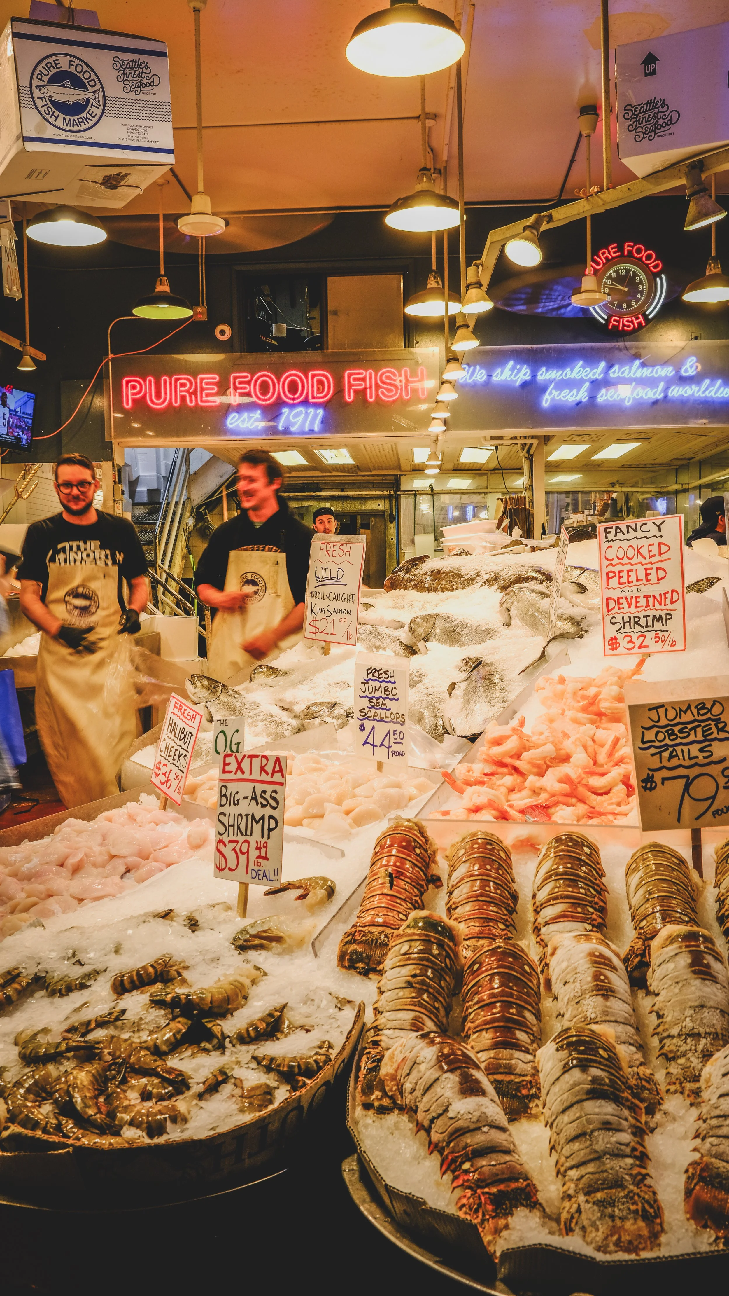 Display of fresh seafood including lobsters, shrimp, and scallops at a fish market with bright signs and staff serving customers.