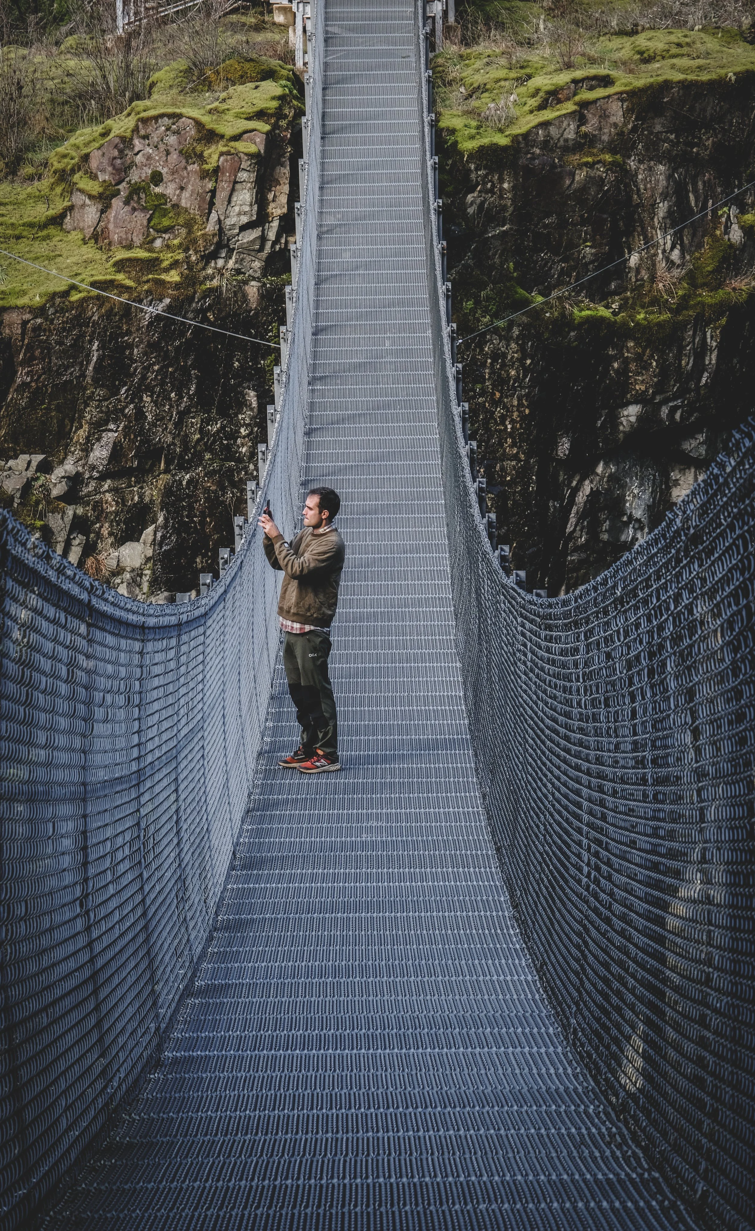 A person standing on a long suspension footbridge taking a photo with a smartphone, with rocky, moss-covered cliffs and green vegetation in the background.